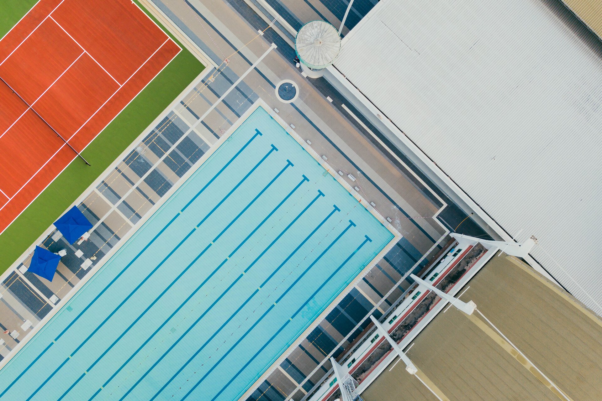 Zdjęcie przedstawia ośrodek sportowy z lotu ptaka. Na środku zdjęcia widać basen z kilkoma torami do pływania, a po lewej stronie kort tenisowy.