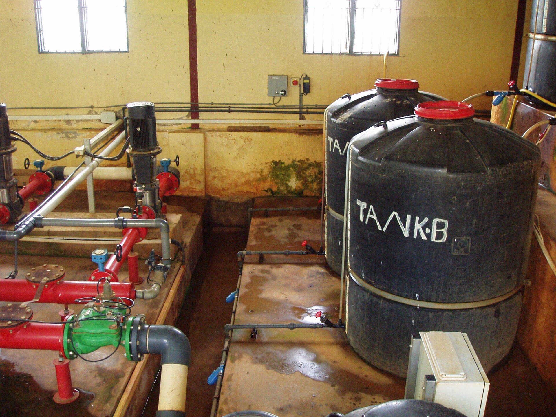 Zdjęcie przedstawia halę i osprzęt służący do mieszania nawozu z wodą. Po lewej stronie widać ciąg rur, a po prawej dwa duże czarne baniaki z czerwonymi zakrętkami. Na wprost widoczna jest ściana z dwoma oknami.