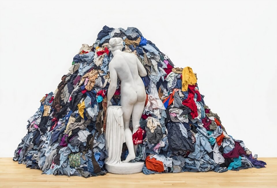 Zdjęcie ukazuje posąg nagiej Wenus odwróconej tyłem i zagłębionej w wysokiej stercie kolorowych szmat.  Szmaty układają się w wysoką piramidę sięgającą powyżej głowy posągu.