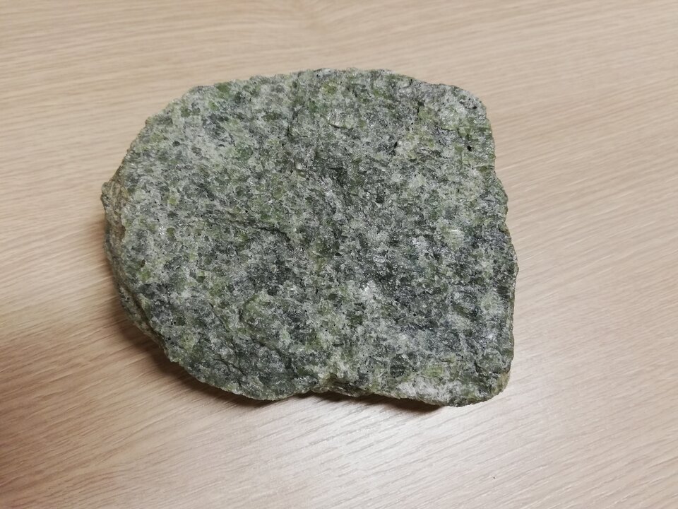 Zdjęcie przedstawia perydotyt. Posiada chropowatą, nierówną powierzchnię o szaro‑zielono‑białej barwie.