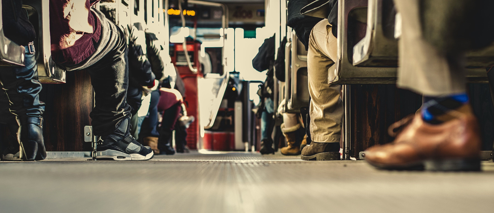 Zdjęcie przedstawia widok z poziomu podłogi autobusu na siedzących w nim ludzi. Widoczne jest przejście, po bokach którego znajdują się siedzący pasażerowie ukazani od dołu. Widoczne są ich nogi i buty. W tle widoczna jest niewyraźnie kabina kierowcy autobusu.
