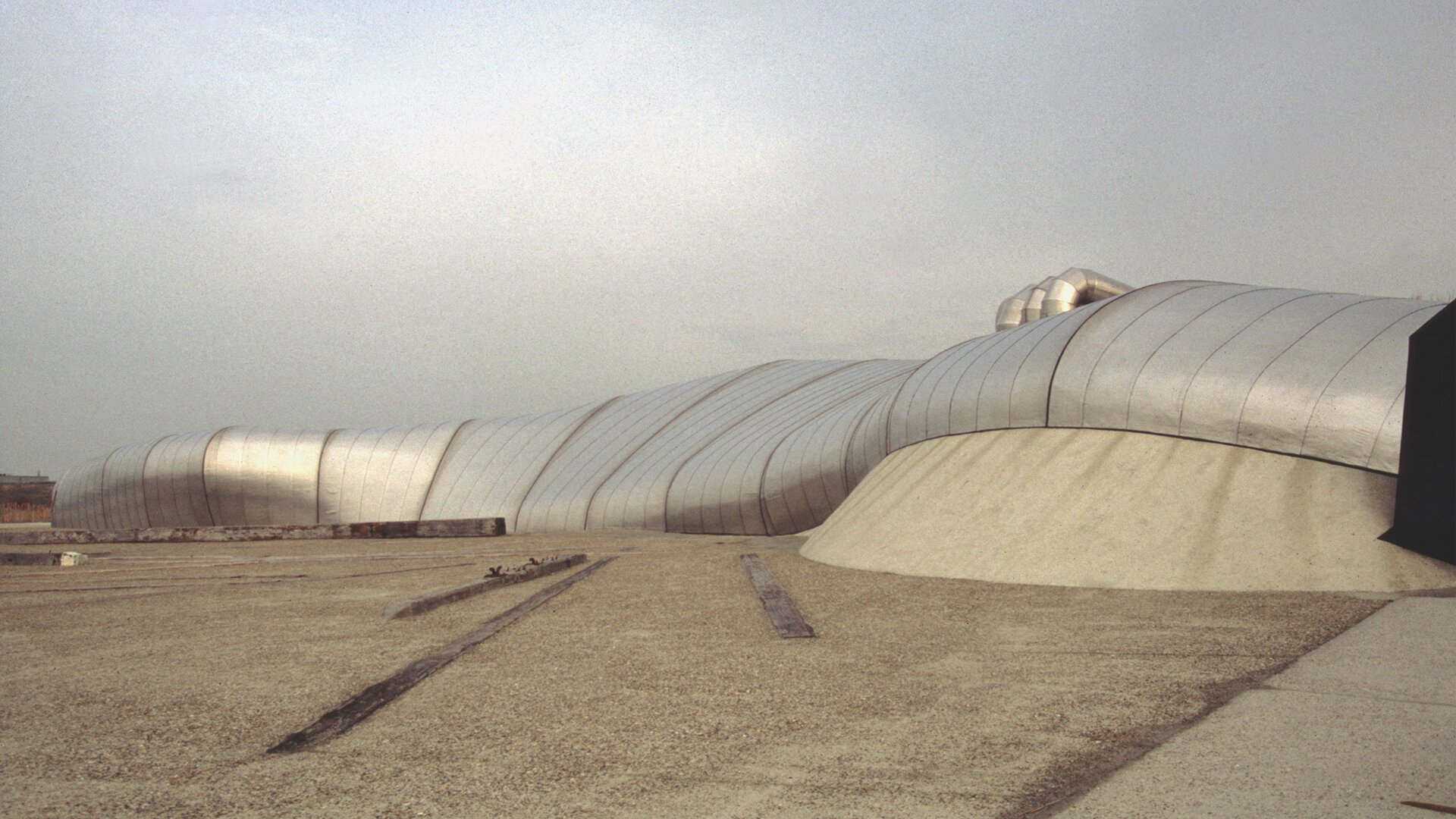 Fotografia przedstawia pawilon „HtwoOexpo” z zewnątrz, na tle szarego nieba oraz brązowej ziemi. Budynek jest długi, wykonany z materiału przypominającego odbijający światło słoneczne metal. Obecne są liczne wybrzuszenia oraz spłaszczenia, nadające jego powierzchni płynności. Konstrukcja jest długa, płaska oraz jednolita - pozbawiona oddzielnych elementów dachu, ścian oraz podstawy.