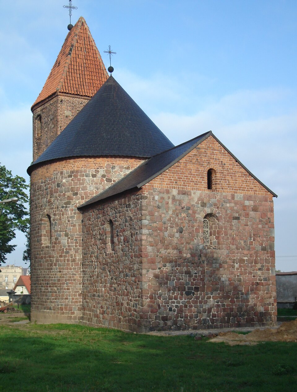 Ilustracja o kształcie pionowego prostokąta przedstawia fotografię rotundy św. Prokopa w Strzelnie. Budowla składa się z brył geometrycznych: walca, stanowiącego nawę, kwadratowego prezbiterium i kwadratowej wieży. Otoczona jest trawą. W tle widoczne są budynki mieszkalne i błękitne niebo.  