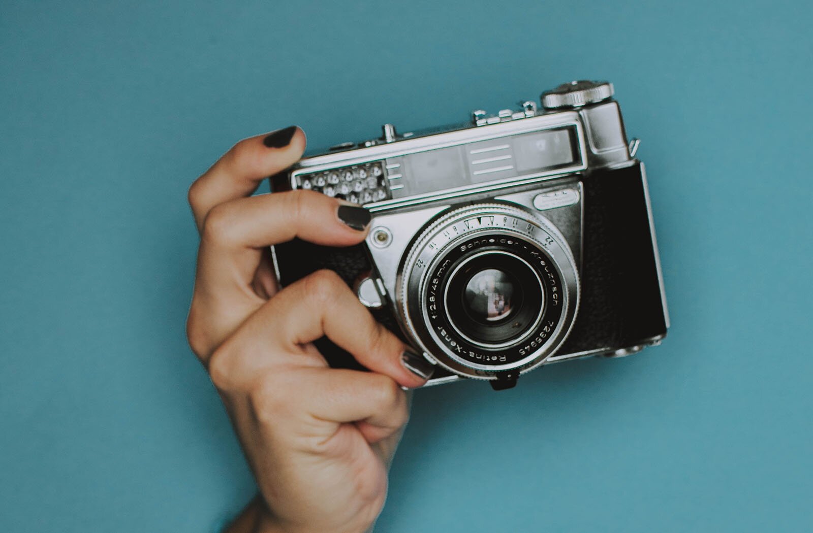 Zdjęcie przedstawia prawą dłoń trzymającą mały aparat analogowy zwrócony obiektywem na  wprost.