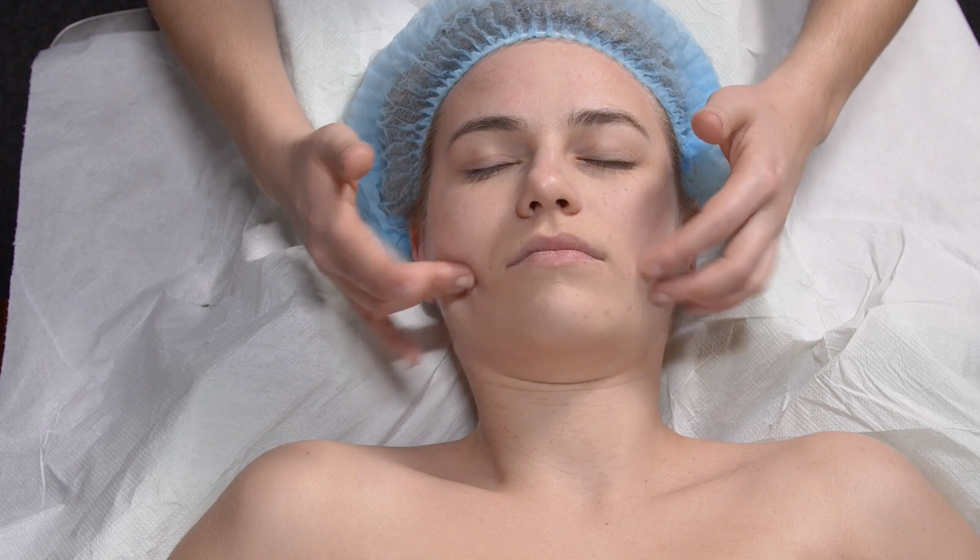 Zdjęcie przedstawia twarz pacjenta w leżeniu tyłem. Głowa pacjenta spoczywa na poduszce. Masażysta znajduje się za głową pacjenta i wykonuje oklepywanie opuszkami wszystkich palców obu rąk. Oklepuje dekolt, szyję, policzki wokół oczu, skronie i czoło.