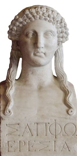 Ilustracja przedstawia rzeźbę – wykonane z białego marmuru popiersie kobiety o długich, opadających na ramiona włosach. Jej głowę zdobi opaska złożona z trzech rzędów równych, małych kwiatów. Na piersi kobiety widnieje napis w języku greckim. Tło jest białe.