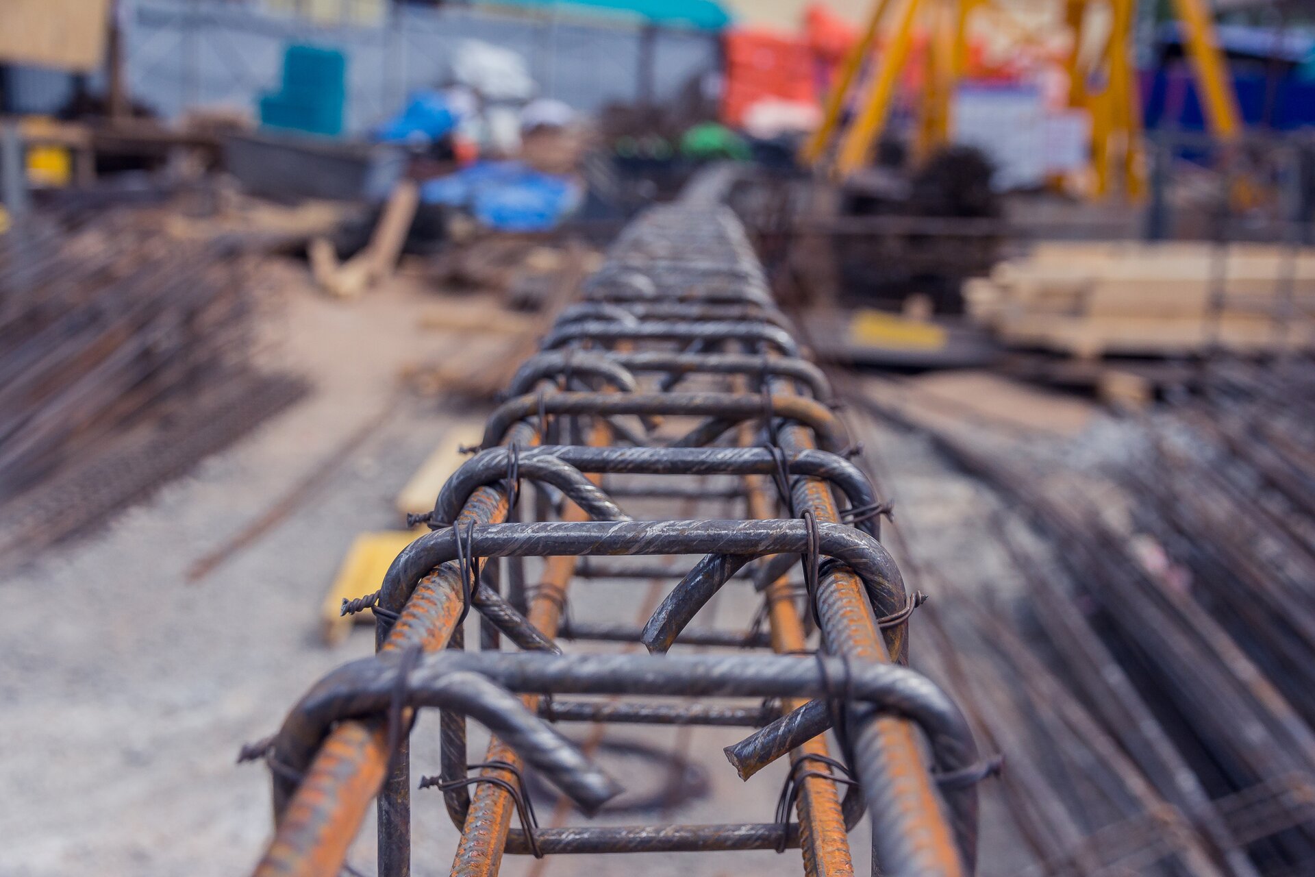 Zdjęcie przedstawia stal zbrojeniową, która jest widoczna na pierwszym planie. W zamazanym tle widać niewyraźnie dwa stosy prętów bo obydwu stronach konstrukcji.