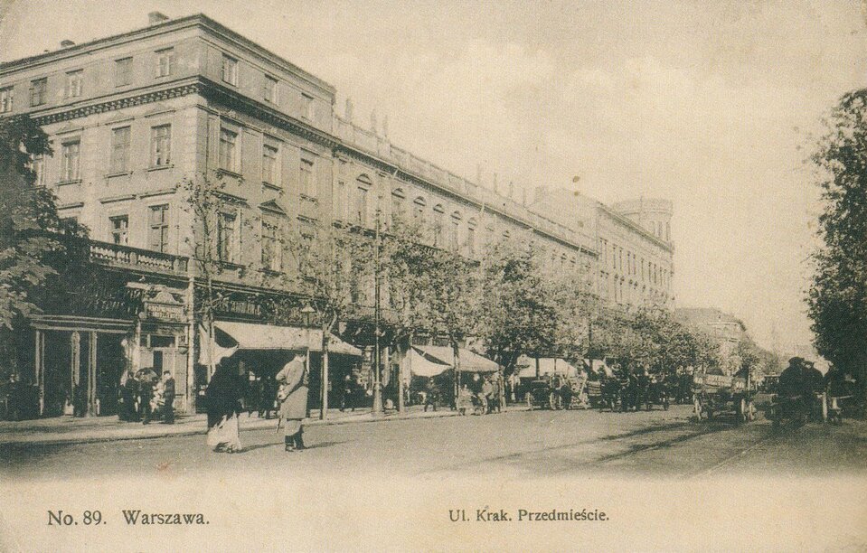 Zdjęcie przedstawia szeroką aleję, biegnącą między drzewami. Po chodnikach przy alei chodzą ludzie. Ulicą jadą dorożki. Przy ulicy stoją duże kamienice, na parterze rozłożone są markizy osłaniające chodnik przed kamienicą.