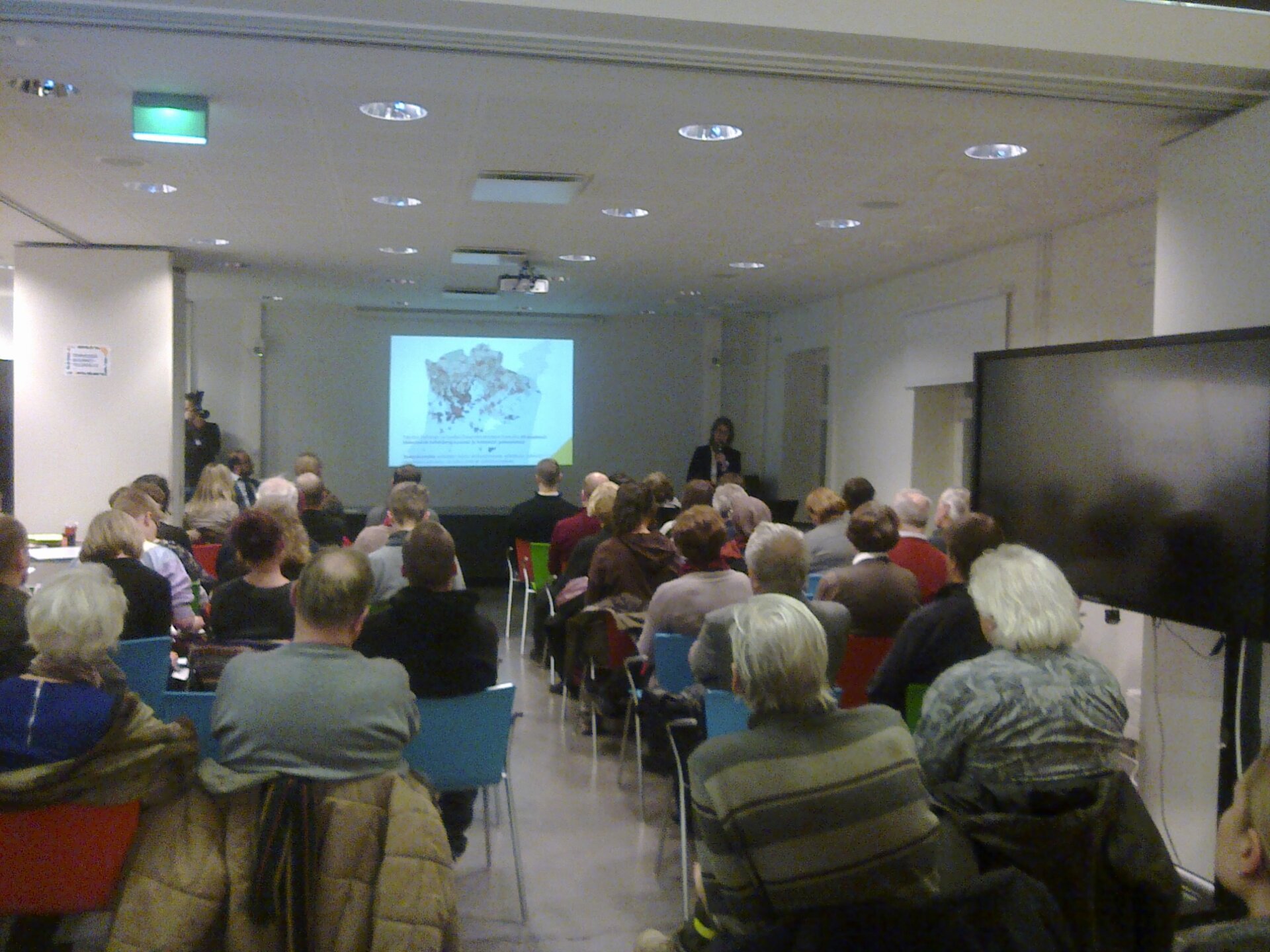 Zdjęcie przedstawia salę, w której odbywa się prezentacja. Na krzesłach siedzą słuchacze w różnym wieku. Patrzą na slajdy wyświetlane na ścianie.