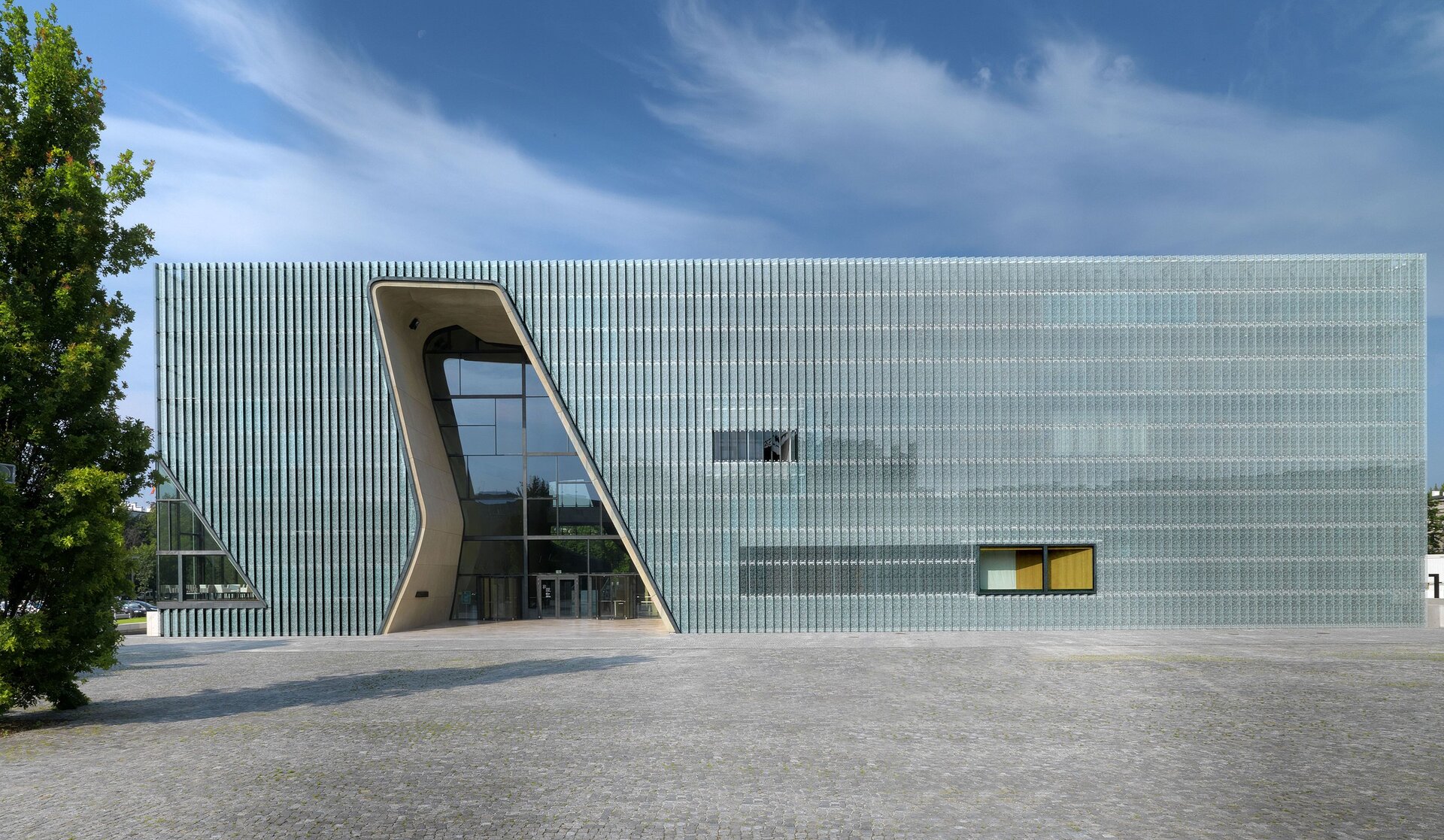 Ilustracja przedstawia Muzeum Żydów Polskich w Warszawie. Na ilustracji znajduje się budynek o prostokątnym kształcie, a jego zewnętrzna część składa się ze szklanych paneli. Główne wejście do budynku przedstawia hebrajską literę law. W tle budynku znajdują się drzewo oraz błękitne niebo.