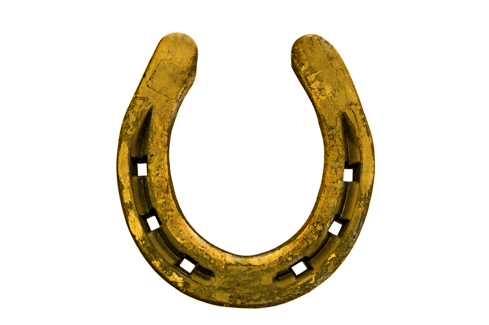Ilustracja przedstawia złotą podkowę, widoczną na białym tle. Podkowa ma klasyczny kształt litery „U” z symetrycznymi ramionami. Na jej powierzchni widać kwadratowe otwory, przez które standardowo wbija się gwoździe, mocujące ją do kopyta konia. Powierzchnia podkowy nosi ślady zużycia i przetarć.