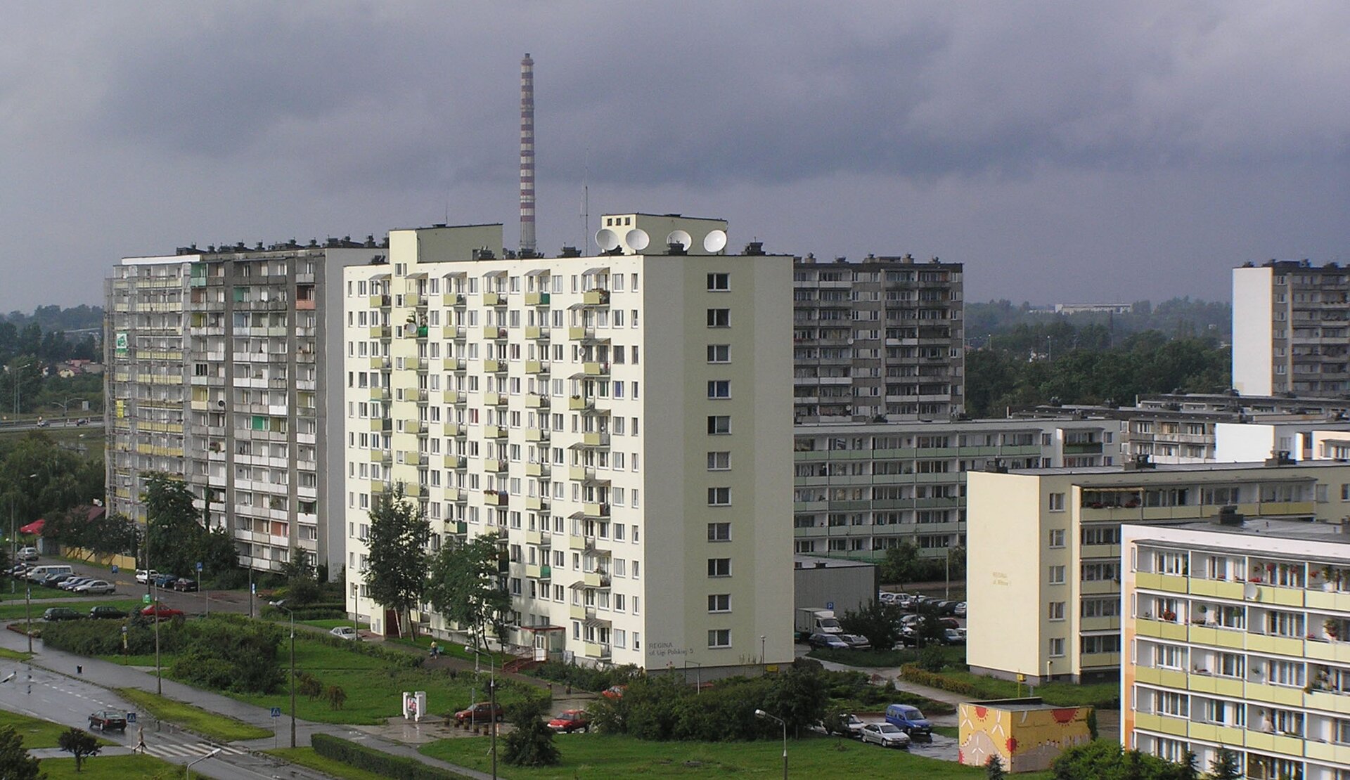 Fotografia przedstawia widok na osiedle mieszkaniowe „Skarpa”, które znajduje się w Toruniu. Na fotografii widoczne są różne bloki, po lewej stronie znajdują się wysokościowce (dziesięciopiętrowe), a po prawej zwykłe, czteropiętrowe. Są one w kolorach jasnoszarym oraz kremowym, widoczne są liczne balkony oraz okna. Bloki są w kształcie prostokątów. Obok bloków oraz naprzeciwko po lewej stronie widoczne są drogi dojazdowe oraz drzewa. W tle widać zazielenione drzewa, samochody – zaparkowane oraz znajdujące się na drodze, szare niebo oraz pobliskie budynki. Fotografia została wykonana w zachmurzony, deszczowy dzień.