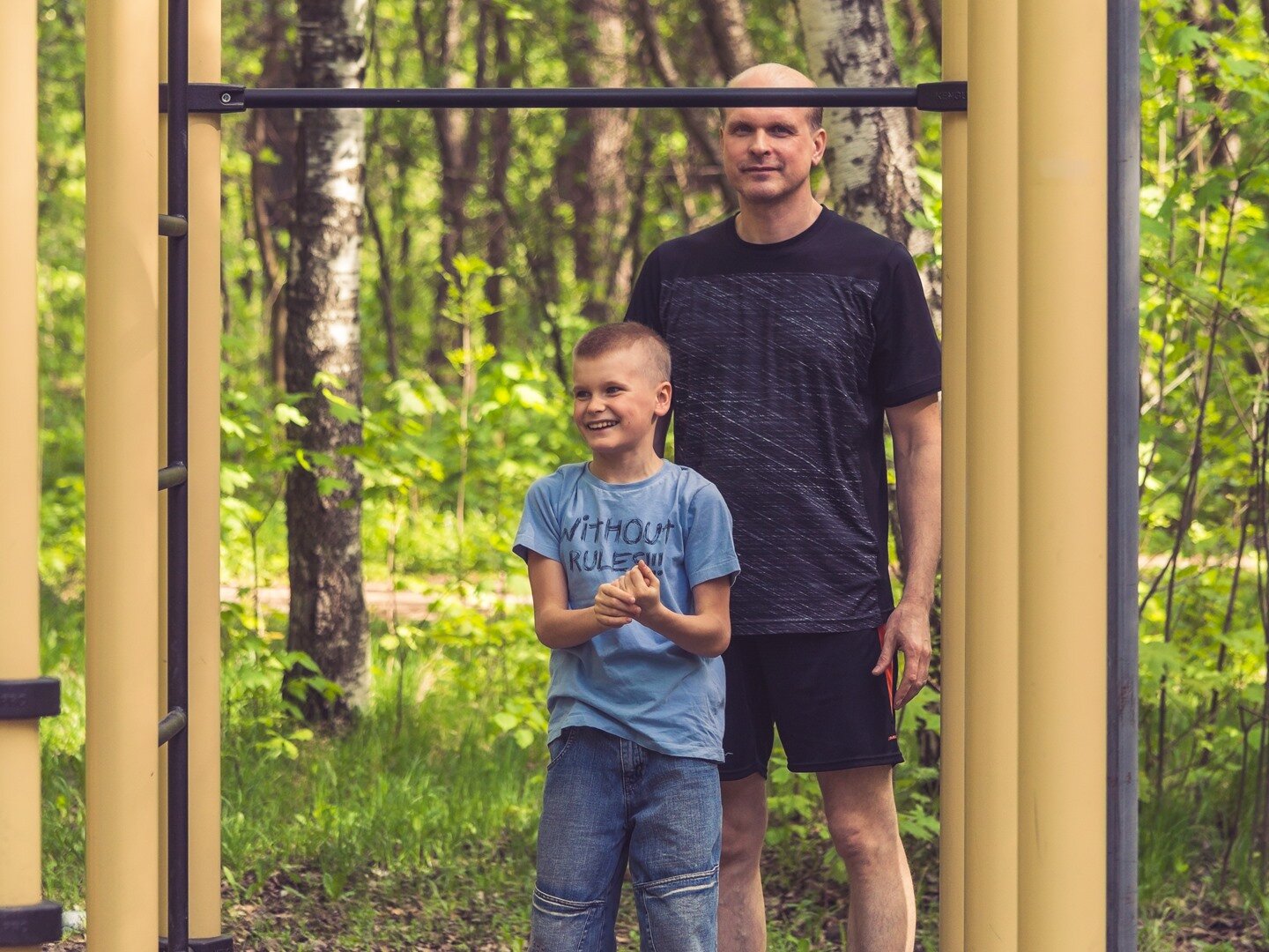 Zdjęcie przedstawia zewnętrzną siłownię, wokoło której rośnie wiele drzew. Przy słupach połączonych drążkiem stoi mężczyzna w średnim wieku w letnim stroju sportowym, a przed nim uśmiechnięty chłopiec. 