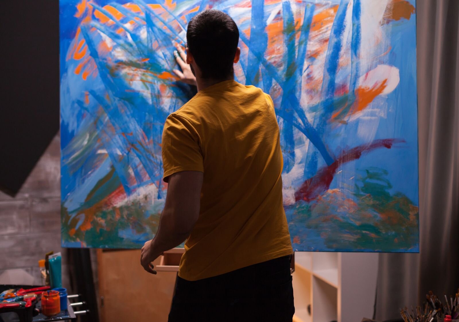 Na zdjęciu przedstawiona jest pracownia i postać malarza przed płótnem obrazu odwrócona tyłem. Mężczyzna maluje palcami, zamaszystymi ruchami, na obrazie powstaje ekspresyjna abstrakcja.