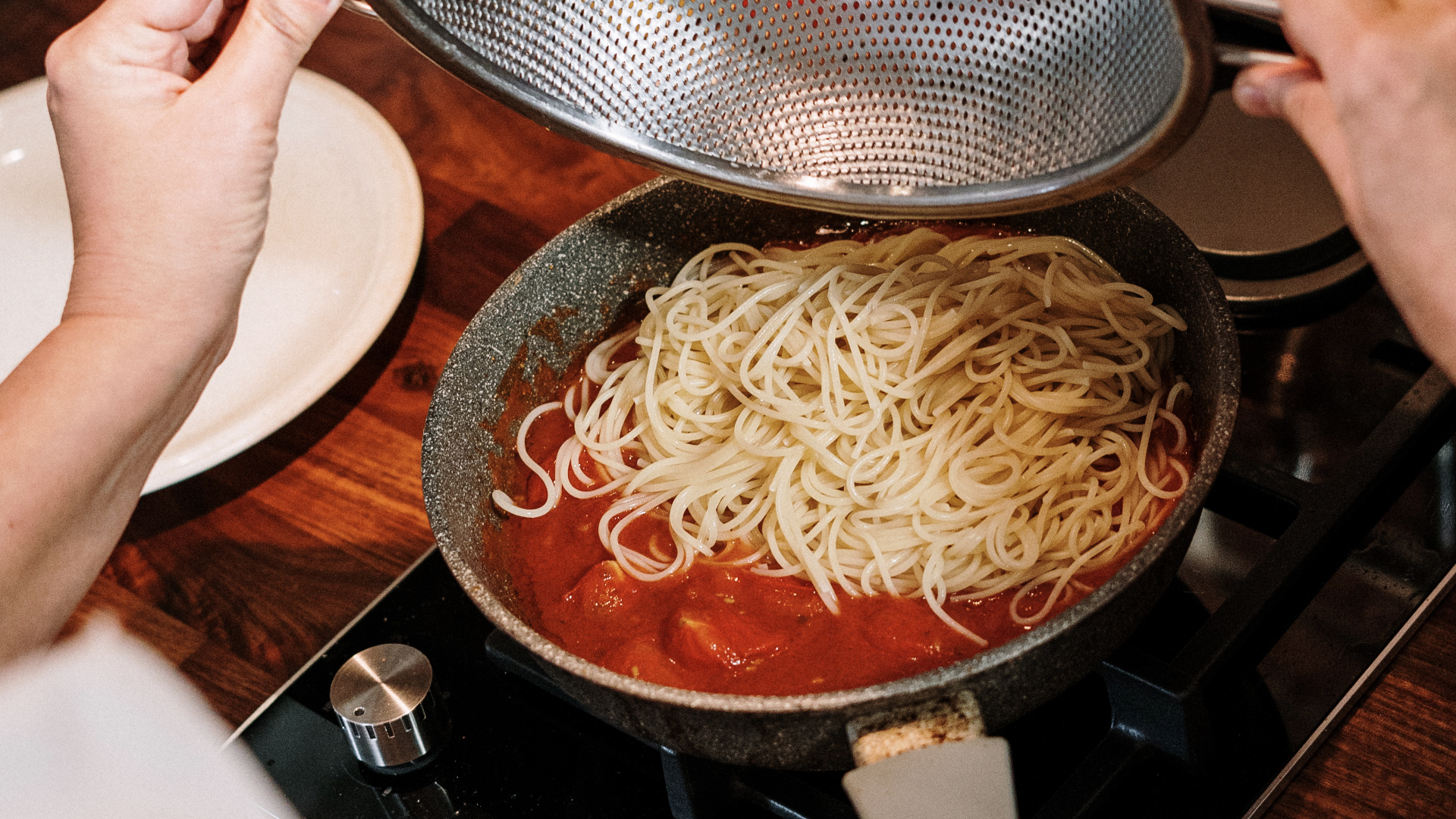 Zdjęcie przedstawia garnek stojący na kuchence. Widoczny jest makaron dodany z durszlaka do sosu pomidorowego.