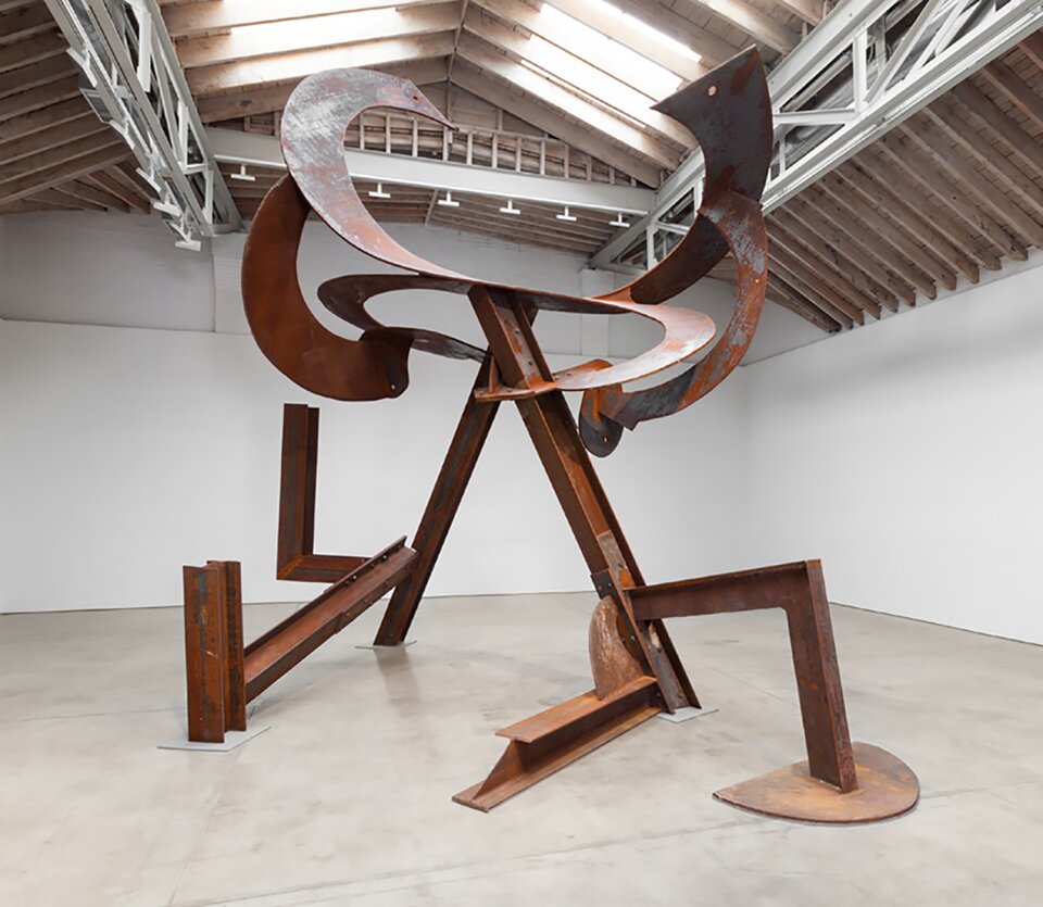 Fotografia przedstawia monumentalną instalację artystyczną umieszczoną w jasnym pomieszczeniu o wysokim suficie. Dzieło składa się z połączonych ze sobą metalowych elementów, przypominających miedziane bądź pokryte rdzą. Elementy przypominają grube blaszki, połączone zawiasami. Znajdują się części pionowe i poziome, wspierające konstrukcję. Przeważają figury geometryczne – prostopadłościany i półokręgi. U góry umieszczono kilka elementów łukowych, swoim kształtem przypominające sierpy. Kompozycja instalacji jest dynamiczna i otwarta.