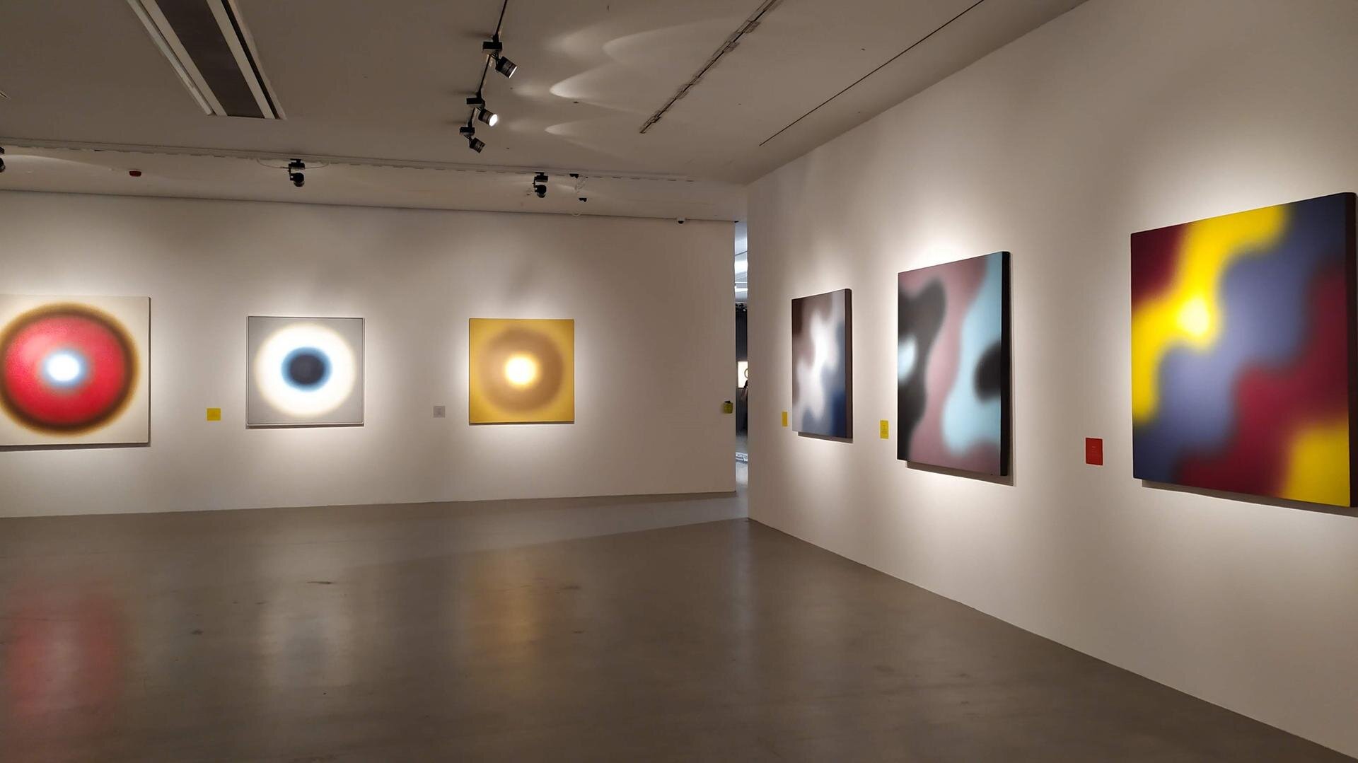 Zdjęcie przedstawia fragment wystawy. Wystawa została urządzona w dużym jasnym pomieszczeniu, gdzie każdy z obrazów ma własne oświetlenie umieszczone pod płótnem. Na zdjęciu znajduje się sześć prac autora. Trzy z nich przedstawiają kolorowe okręgi, a trzy kolorowe plamy. 