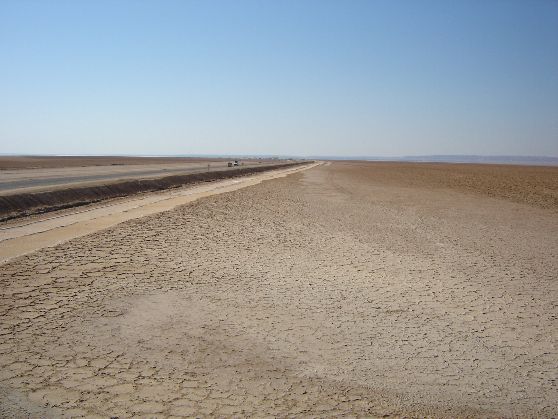Na fotografii znajduje się Szott, bezodpływowe, słone jezioro w Tunezji. Fotografia przedstawia fragment wyschniętego dna jeziora. Po horyzont prowadzi droga, a obok niej biegnie szeroki pas spękanej ziemi.