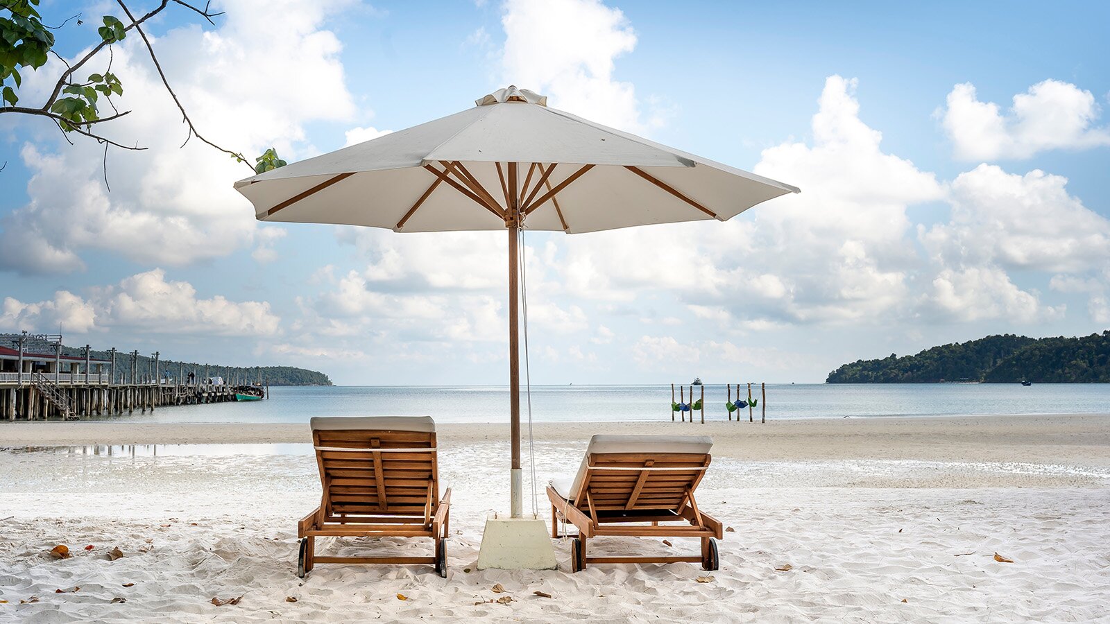 Zdjęcie ukazuje piaszczystą plażę u wybrzeży morza lub oceanu. Na plaży stoją dwa drewniane leżaki, oraz pasujący do nich duży beżowy parasol. Piasek na plaży jest biały.
