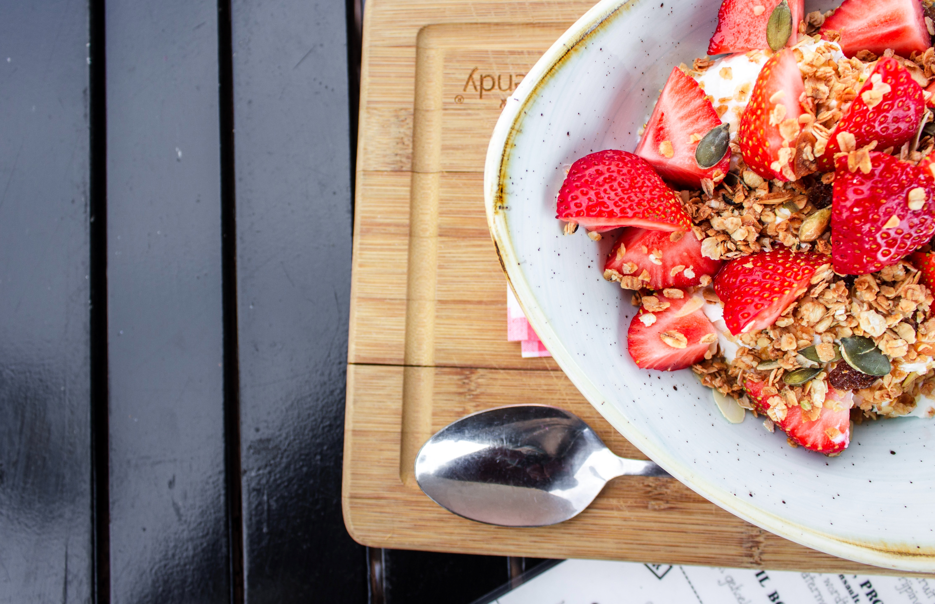 Zdjęcie przedstawia owsiankę z dużą ilością truskawek. Owsianka znajduje się na talerzu. Talerz stoi na drewnianej desce. Obok talerza leży łyżka.