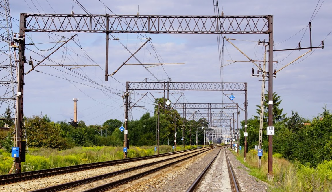 Zdjęcie przedstawia bramkę trakcyjną. na zdjęciu widać trzy tory kolejowe, wzdłuż których ustawione są bramki trakcyjne. Oparte są one na podporach stojących przy zewnętrznych krawędziach torów. Między podporami rozwieszone są dźwigary, pod którymi zawieszone są wsporniki z podwieszonymi sieciami górnymi dla wszystkich torów.