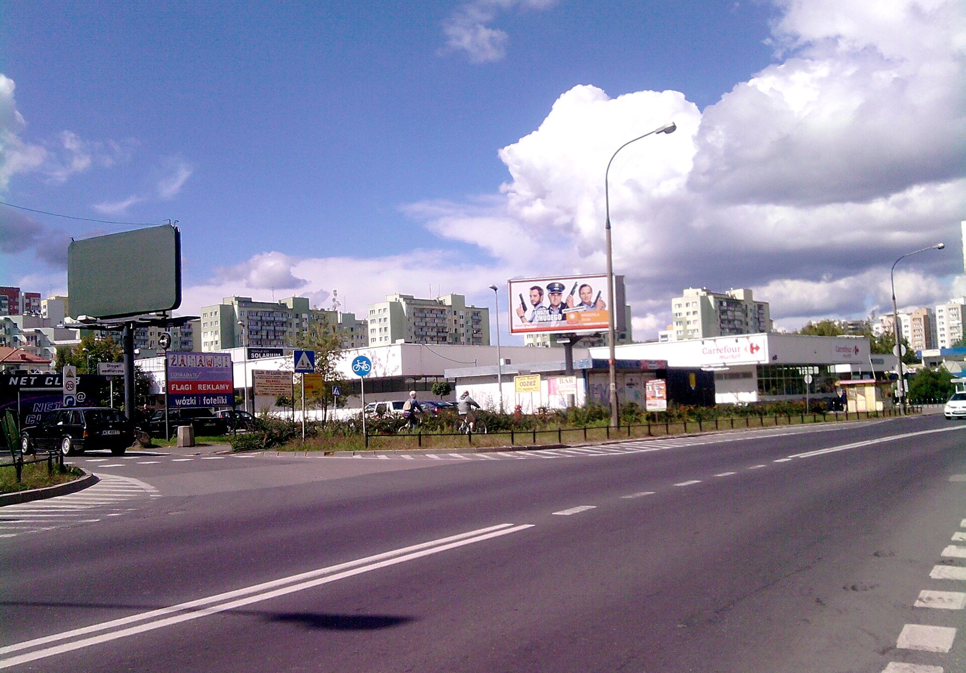 Zdjęcie przedstawia ulicę, wzdłuż której znajduje się kilka tablic reklamowych. Za nimi jest płaski, duży budynek marketu. W oddali osiedle blokowisk. 