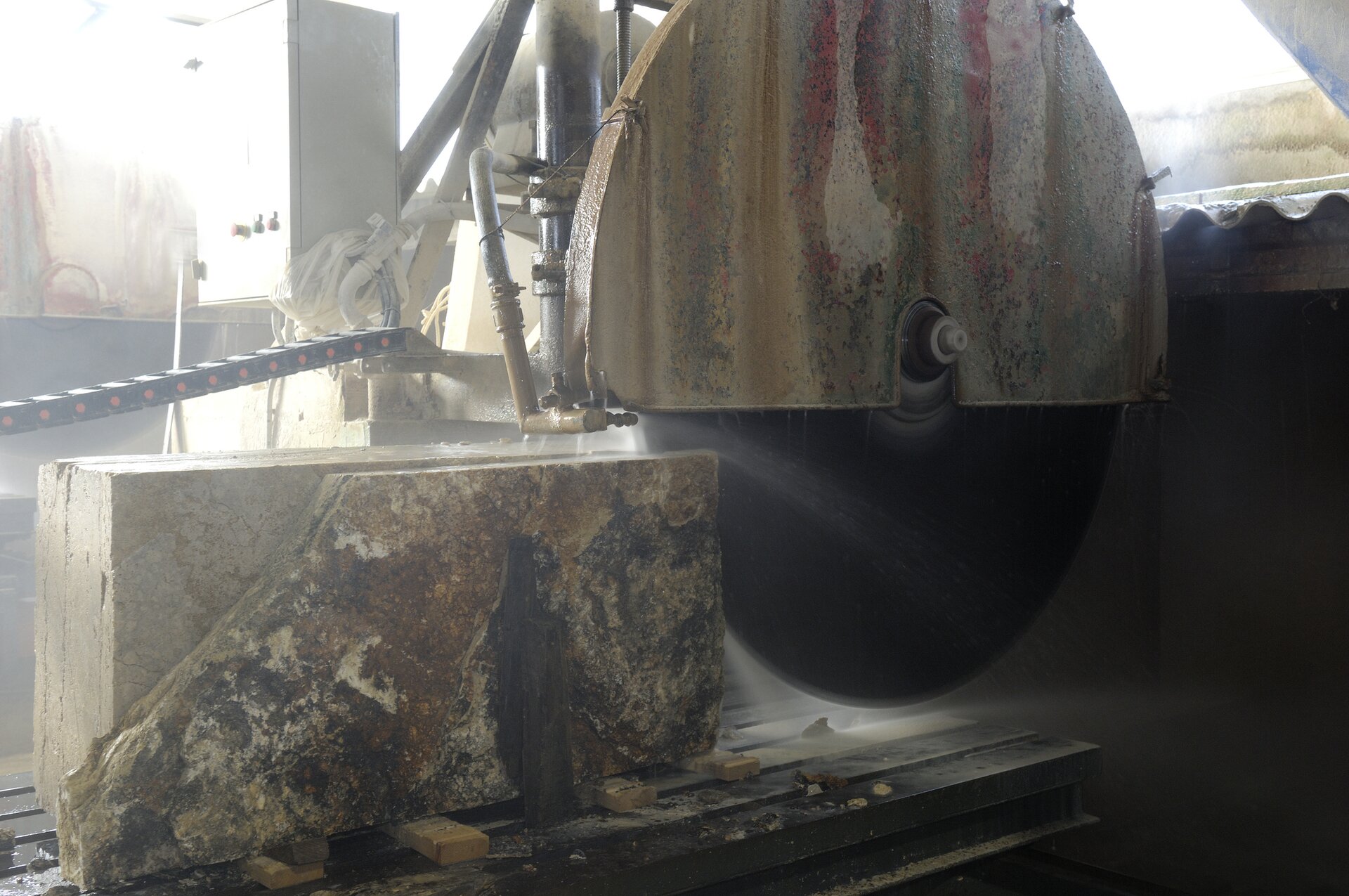 Zdjęcie ukazuje cięcie bloku za pomocą traka tarczowego. Zawieszona na ramie tarcza tnąca zagłębia się w kamienny element, rozcinając go. Na kamieniu oraz na obudowie piły widoczne są krople wody.