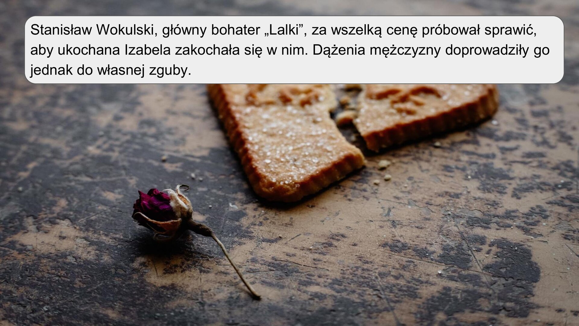 Zdjęcie przedstawia ciastko maślane w kształcie serca, które zostało złamane. Na ciastu pojawia się napis “LOVE”. Poniżej leży uschnięty pączek czerwonej róży. Zarówno ciastko, jak i pączek, leżą na drewnianej, zniszczonej powierzchni. Na zdjęciu znajduje się napis: „Stanisław Wokulski, główny bohater „Lalki”, za wszelką cenę próbował sprawić, aby ukochana Izabela zakochała się w nim. Dążenia mężczyzny doprowadziły go jednak do własnej zguby”.