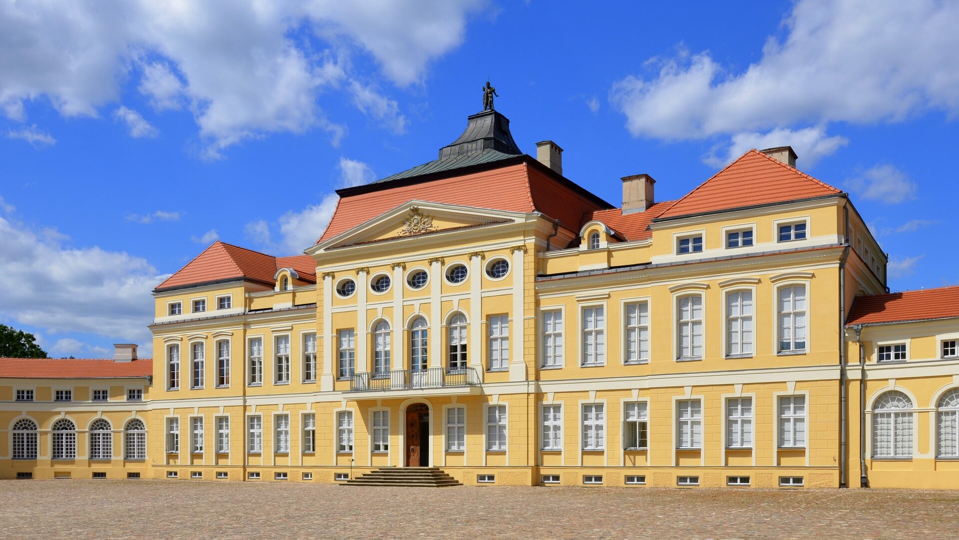 Fotografia przedstawiająca Pałac Raczyńskich w Rogalinie, 1768–1776. Jest to zespół pałacowo‑parkowy zbudowany w stylu późnobarokowym. Prostokątne były budynku wypełnione są bardzo licznymi oknami. Przed wejściem znajdują się niewielkie schody. Nad wejściem balkon. Dach z czerwonej cegły. Na samym szczycie dachu znajduje się rzeźba postaci męskiej. 