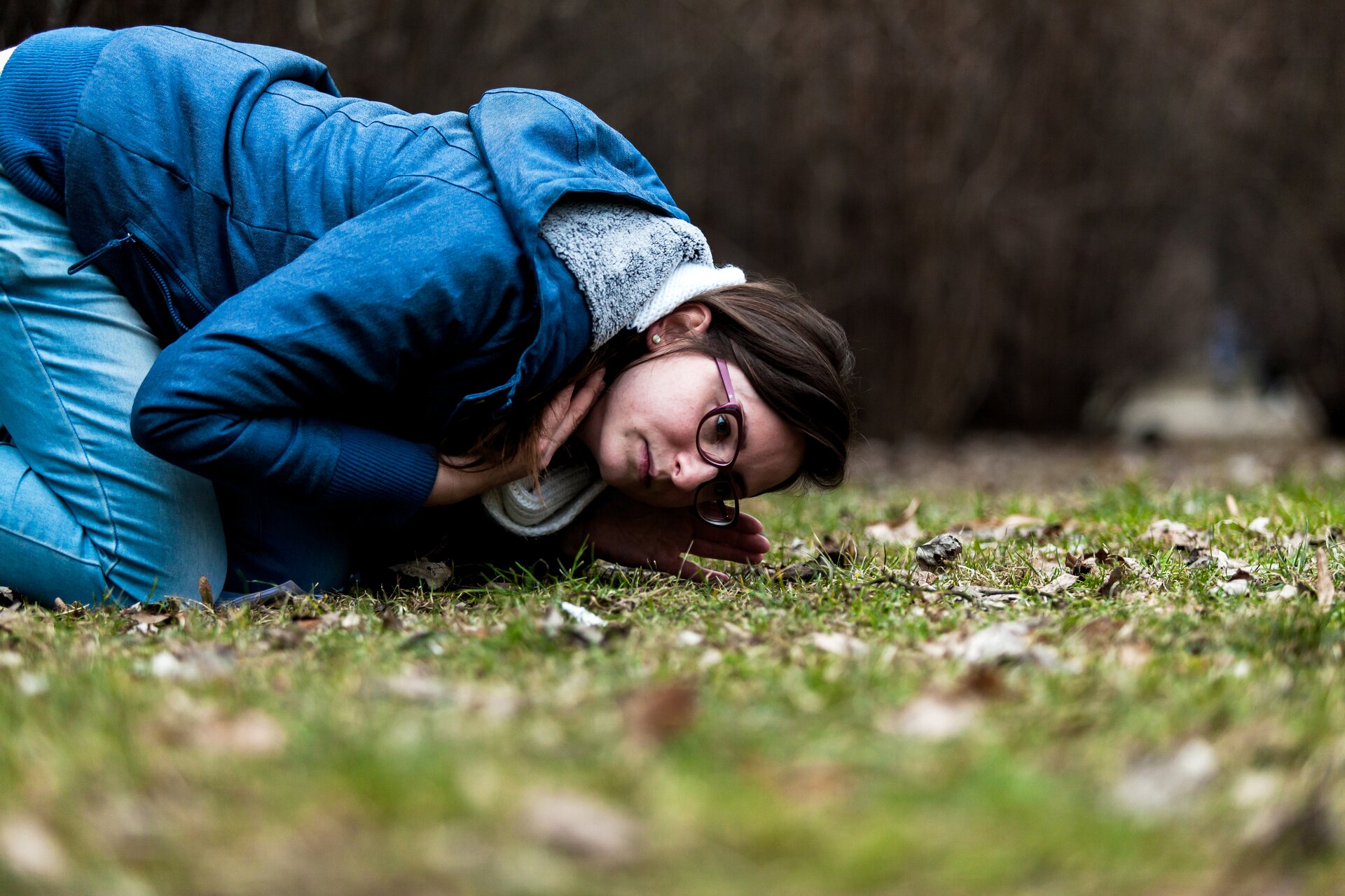 Zdjęcie przedstawia kobietę w granatowym ubraniu i okularach klękającą na ziemi, pochyloną i przykładającą do niej ucho. W tle zieleń.