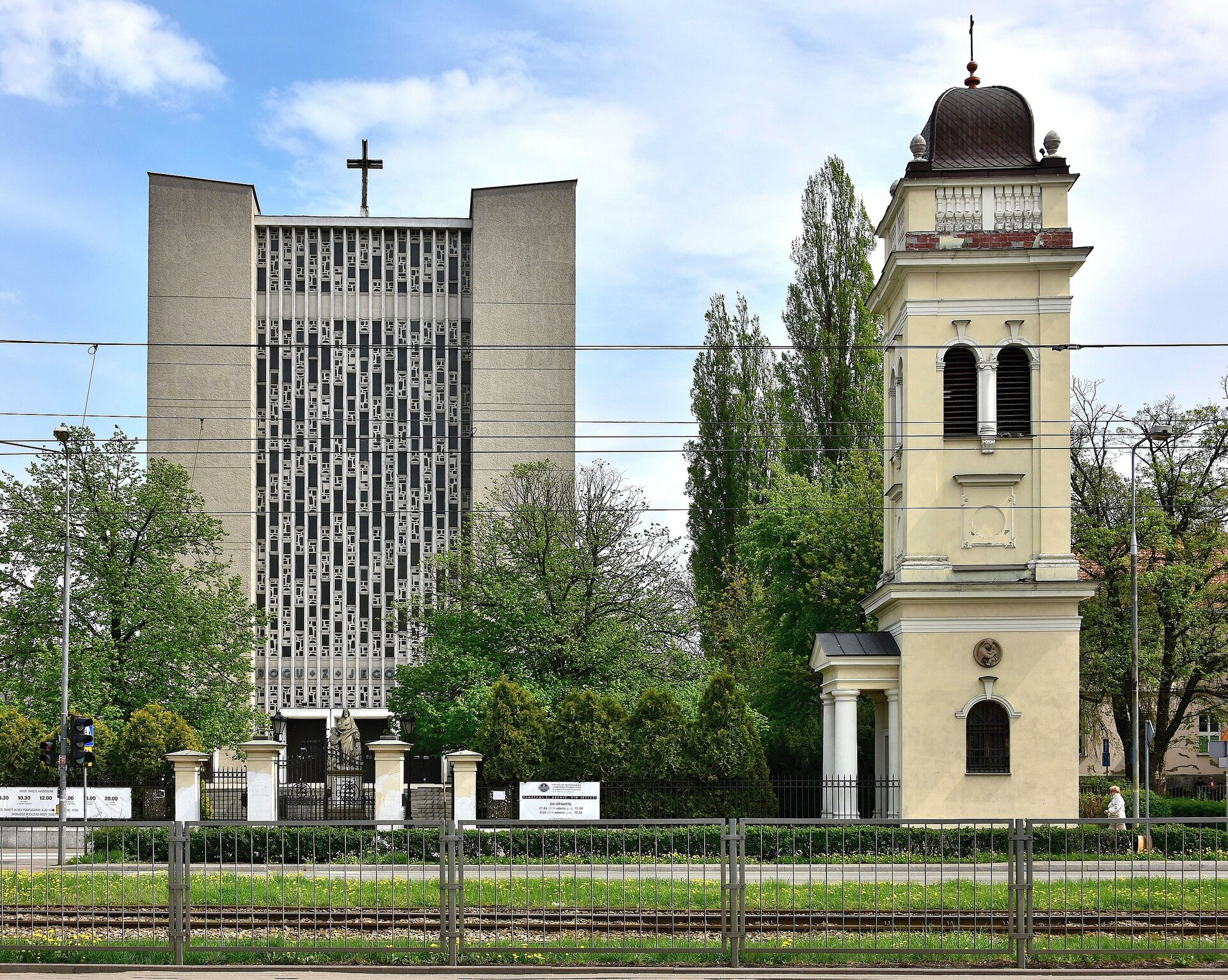 Fotografia przedstawia widok na dwa budynki na tle zachmurzonego, niebieskiego nieba. Pomiędzy oraz przed budynkami znajdują się zielone drzewa. Budynek przedstawiony z lewej strony jest symetryczny, wysoki. Jego część środkową stanowi bryła przypominająca czarny prostokąt, który podzielony jest na mniejsze prostokąty, na którego powierzchni widać jaśniejsze wzory. Po obu jego stronach są gładkie oraz nieco wyższe części budynku, przypominające szare kolumny o skośnych krawędziach górnych. Na dole widać duże wejście, a u góry – krzyż. Budynek po prawej stronie, widoczny bokiem, przypomina wieżyczkę o ozdobnym, ciemnoszarym dachu, na którego wierzchołku również umieszczono krzyż. Na ścianie widoczne są zasłonięte okna, a wejście podtrzymują cztery jasne kolumny. Budynek znajdujący się po lewej stronie to kościół, a budynek znajdujący się po prawej stronie to dzwonnica.