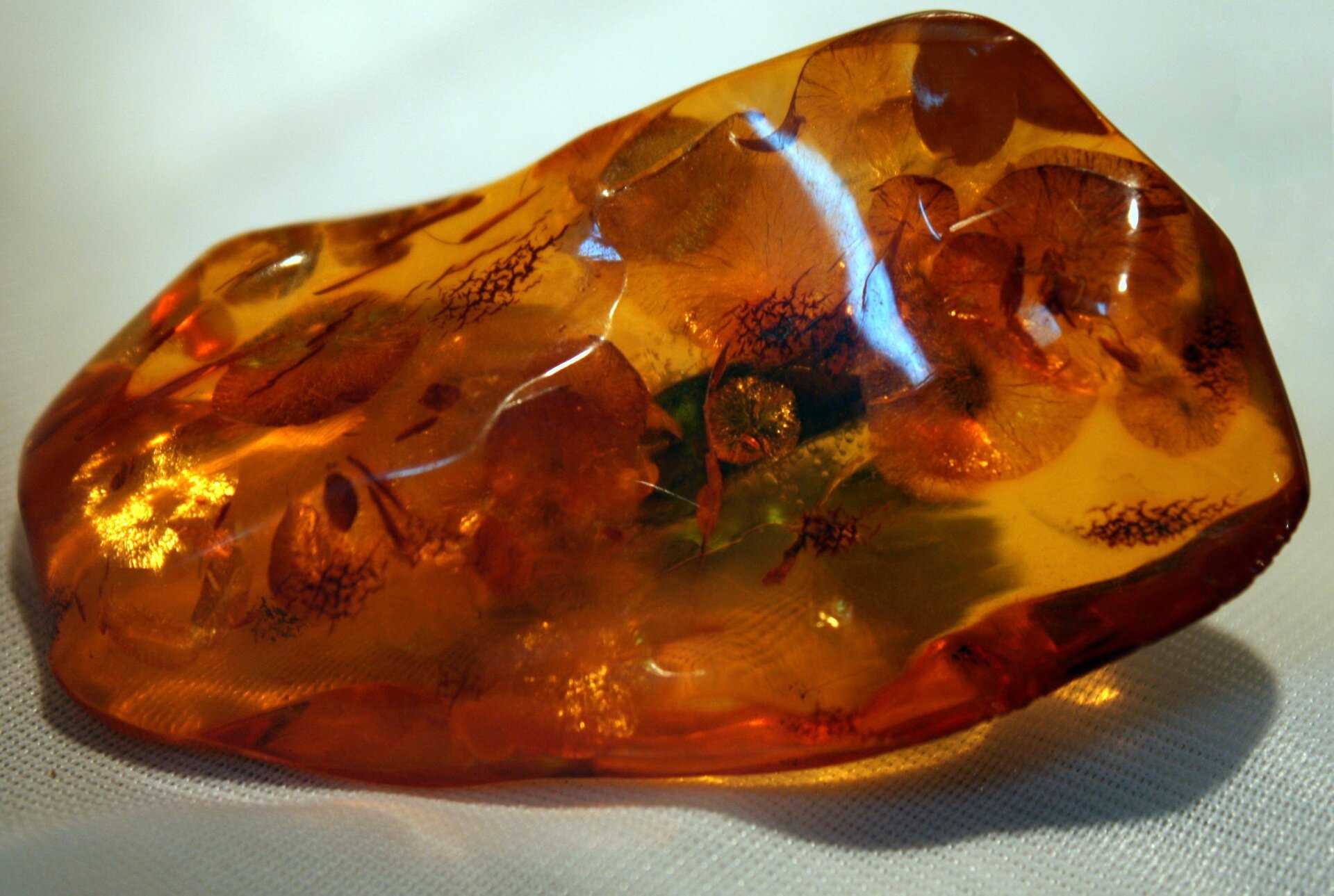 Zdjęcie przedstawia kawałek złocisto czerwonego bursztynu leżącego na skrawku białej tkaniny. Kamień ma nieregularne, zaokrąglone krawędzie i cechuje się dużą przezroczystością.
