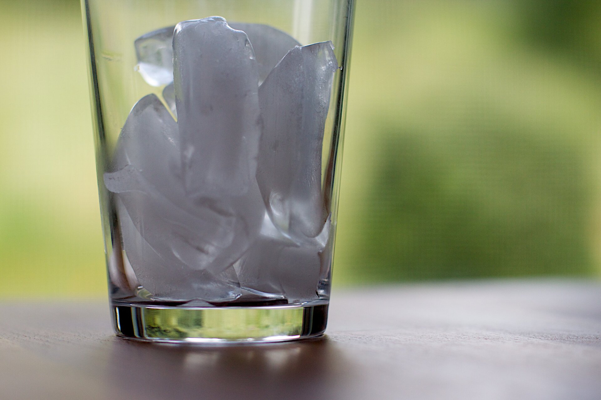 Fotografia przedstawia przezroczystą szklankę z lodem. Wewnątrz szklanki kilka kawałków lodu. Szklanka stoi na stole. Niektóre z kostek topnieją, na dnie widać cienką warstwę wody. Zamazane tło.