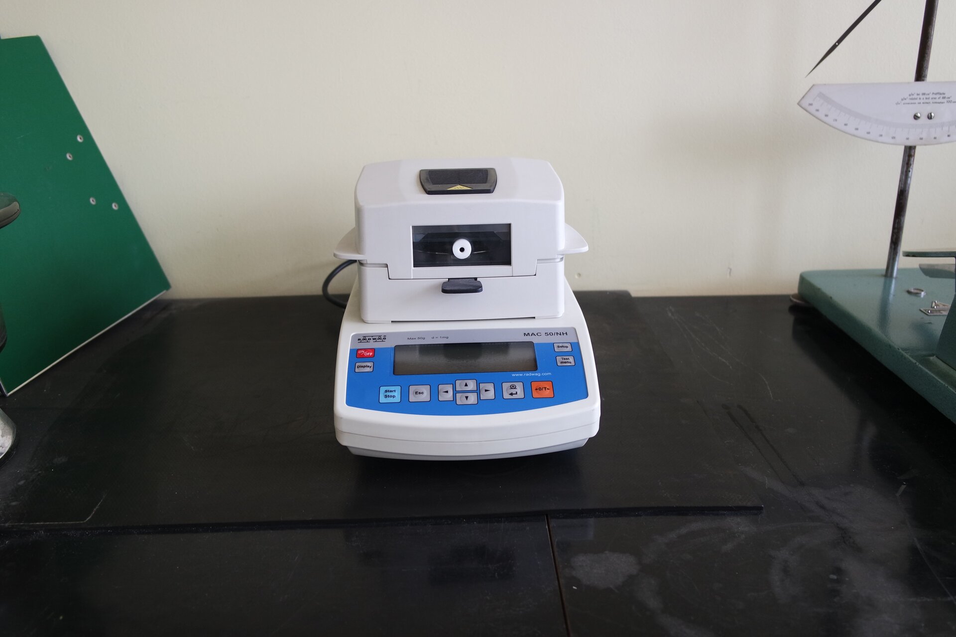 Na zdjęciu ukazano wagosuszarkę do oznaczania wilgotności. Urządzenie zostało umieszczone na blacie. W dolnej jego części widoczny jest wyświetlacz umieszczony na panelu z różnymi przyciskami. Powyżej znajduje się waga oraz komora suszarki.