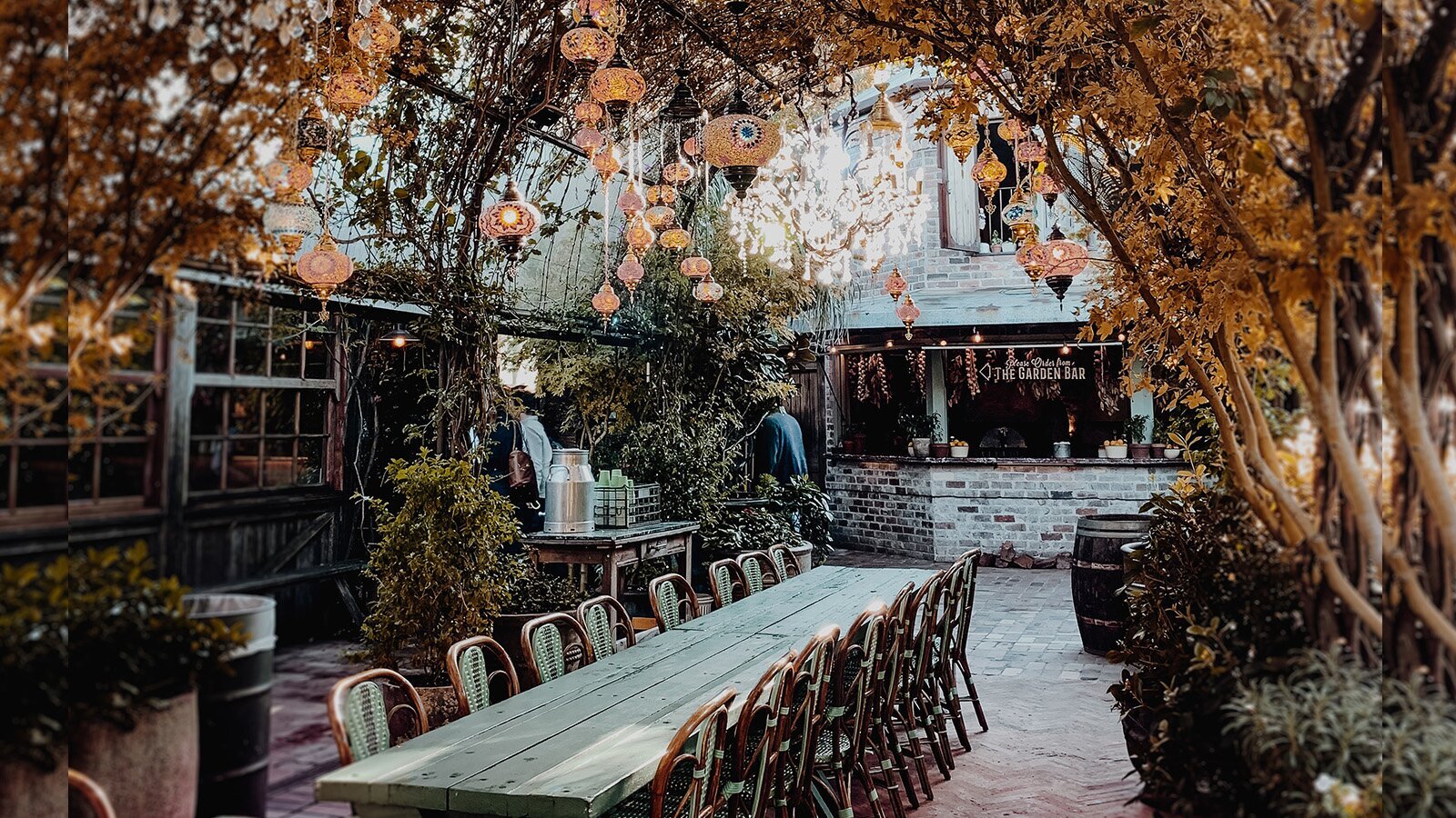 Zdjęcie przedstawia udekorowane patio, na którym widoczny jest długi, drewniany stół z krzesłami ustawionymi wzdłuż dłuższych boków. Po obu stronach stołu widoczne są dekoracyjne lampiony oraz roślinność. Nad całością rozpięta jest łukowata pergola, po której pną się rośliny, tworząc naturalne zadaszenie nad stołem. W tle widoczna jest ceglana lada, nad którą widoczny jest napis w języku angielskim: THE GARDEN BAR.