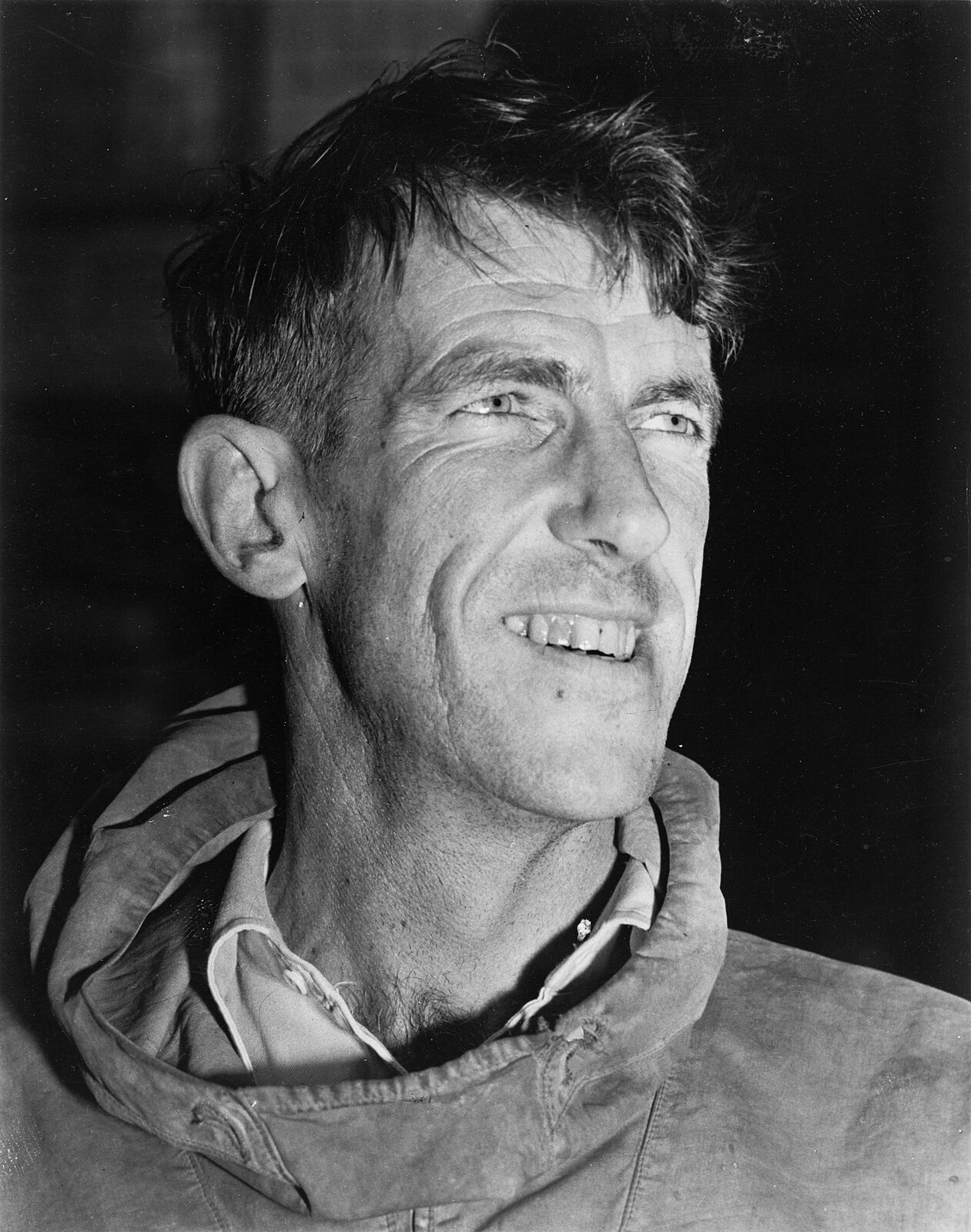 Edmund Hillary (Nowozelandczyk). Zdjęcie jest czarno‑białe. Mężczyzna obrany w bluzę z golfem. Patrzy przed siebie. Jest uśmiechnięty. Nie posiada zarostu, ma krótkie włosy, lekkie zakola, prosty nos, małe oczy, regularne rysy twarzy.