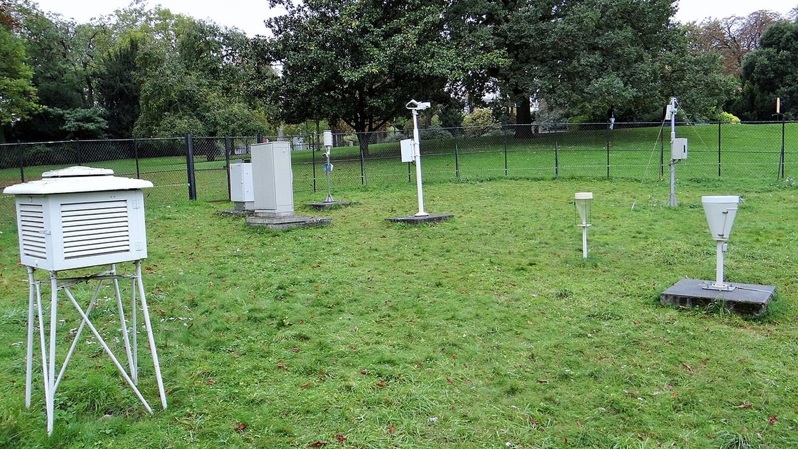 Zdjęcie przedstawia ogródek meteorologiczny. W ogródku widoczne są przyrządy do pomiaru składników pogody, takie jak: deszczomierz, wiatromierz oraz klatka meteorologiczna (z termometrem, barometrem). Ogródek otoczony jest białą siatką.