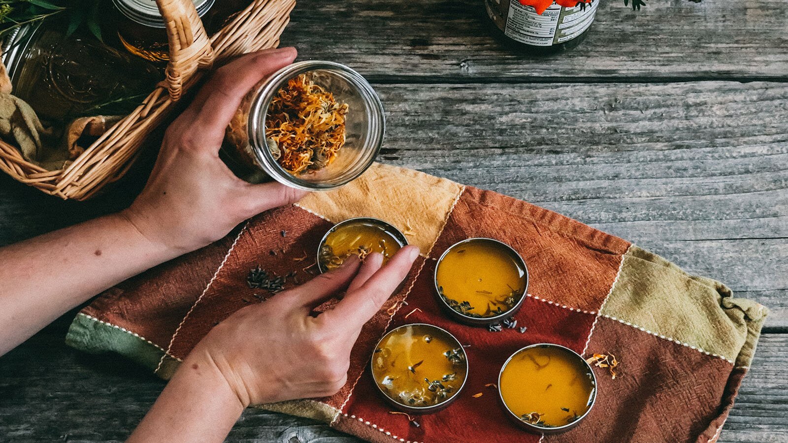 Zdjęcie przedstawia ściereczkę rozłożoną na płaskiej powierzchni, na której ustawiono cztery okrągłe, niskie pojemniczki wypełnione żółto, oleistym płynem. Nad jednym z pojemniczków są dwie ręce. Lewa ręka trzyma słoik wypełniony suszonymi roślinami, prawa ręka, między palcami trzyma trochę mieszanki ze słoika nad pojemniczkami z płynem. 