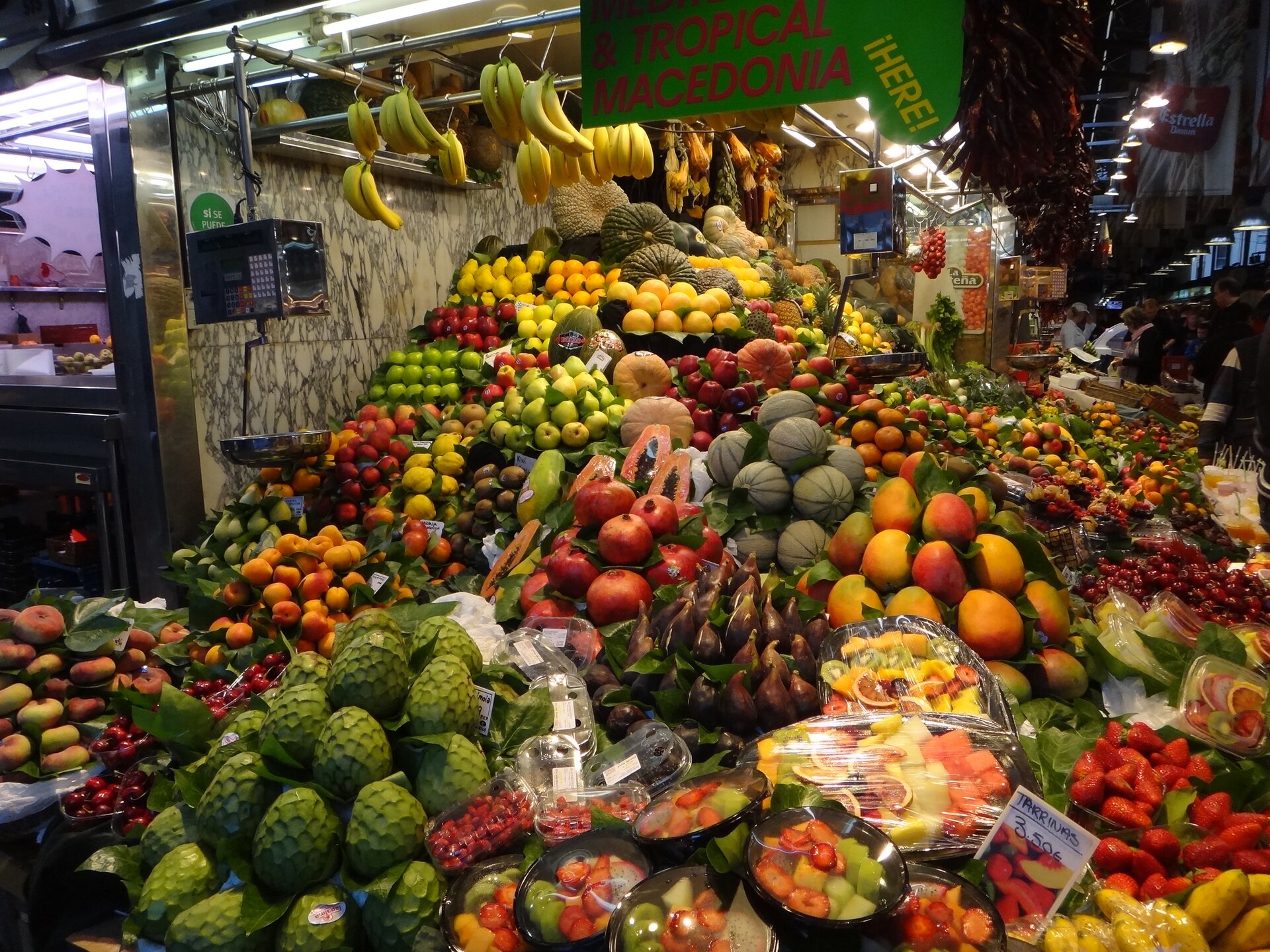 Ilustracja przedstawia świeże owoce rozłożone przed stoiskiem na targu. Na zdjęciu znajdują się między innymi jabłka, truskawki, mango, brzoskwinie i banany.