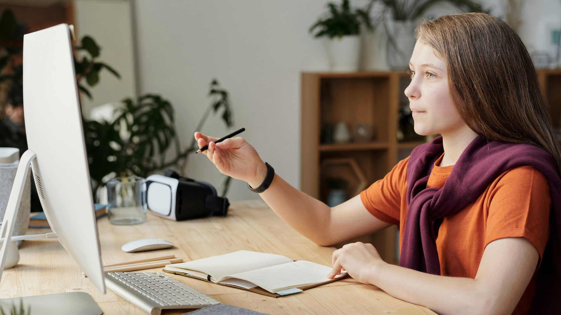 Zdjęcie przedstawia młodą kobietę z długimi włosami siedzącą przed ekranem komputera. Kobieta widoczna jest z boku, w prawej ręce trzyma długopis i unosi ją wskazując w stronę ekranu.