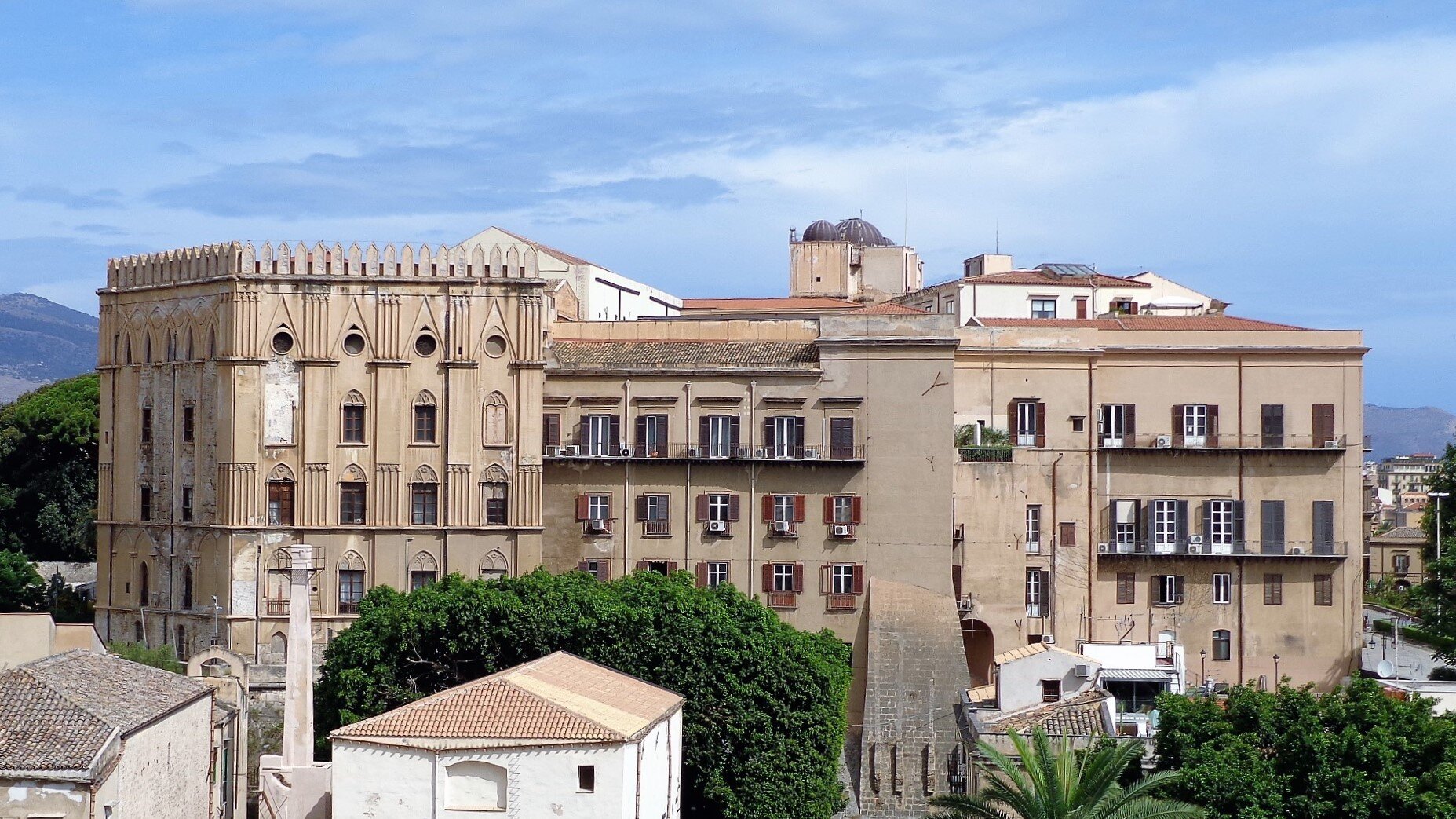 Zdjęcie przedstawia budynek kilkupiętrowego, dużego pałacu. Składa się z rzędów okien oraz brązowej elewacji. Okna na prawej części budynku mają drewniane okiennice. Lewa strona budynku składa się z płaskich kolumn, dekoracji w kształcie ostrych łuków oraz rzędów okien. Wokół stoją niższe budynki. W tle zalesione wzgórza i niebo z chmurami.