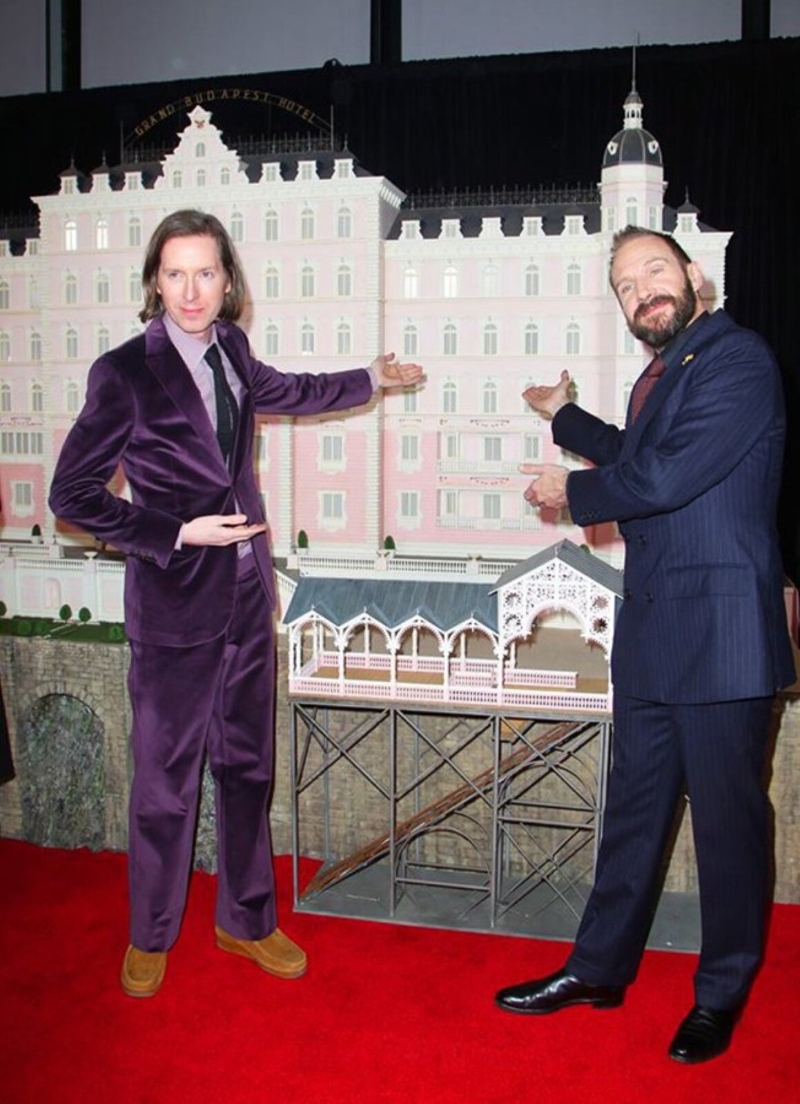 Ilustracja przedstawia makietę Hotelu Grand Budapeszt z lat 30. Na pierwszym planie znajduje się dwóch mężczyzn., którzy stoją na czerwonym dywanie. Mężczyzna po lewej stronie ma dłuższe, ciemne włosy. Ubrany jest w fioletowy garnitur, jasnofioletową koszulę, czarny krawat i brązowe buty. Mężczyzna z prawej strony ma krótkie ciemne włosy, wąsy i brodę. Ubrany jest w granatowy garnitur, czarną koszulę, bordowy krawat i czarne, błyszczące buty. Obaj mężczyźni rękoma wskazują na makietę Hotelu Grand Budapeszt, która znajduje się za nimi. Jest ona w odcieniach różu z białymi oknami i ciemnym dachem. Stoi na konstrukcji z brązowo‑szarej cegły. Przed głównym budynkiem hotelu znajduje się mniejszy budynek z zielonym dachem. Stoi on na metalowej, ażurowej konstrukcji.