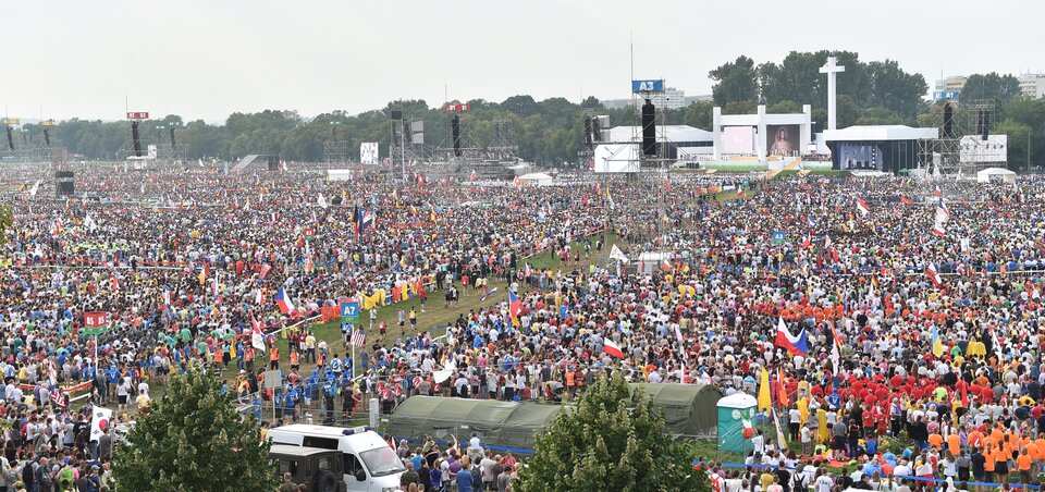 Ilustracja przedstawia fotografię ze Światowych Dni Młodzieży w Krakowie. Wykonane z góry zdjęcie ukazuje tłum ludzi zgromadzonych przed polowym ołtarzem. W tle znajduje się widok na krakowskie Błonie.