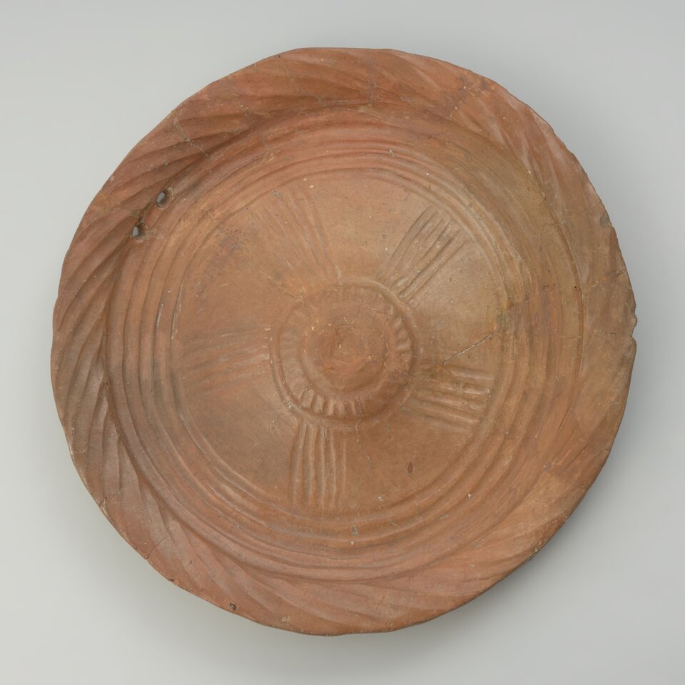 Ilustracja przedstawia zdobiony talerz z okresu epoki brązu, kultury łużyckiej. Na powierzchni talerza widoczny jest wyraźny wzór, który składa się z geometrycznych motywów. W centralnej części znajduje się okrągły, wytłoczony element. Wokół niego rozciągają się symetrycznie spirale i łuki, które tworzą wrażenie dynamicznego ruchu. Brzegi talerza zdobią powtarzające się wytłoczenia w formie prostych linii lub małych punktów. 