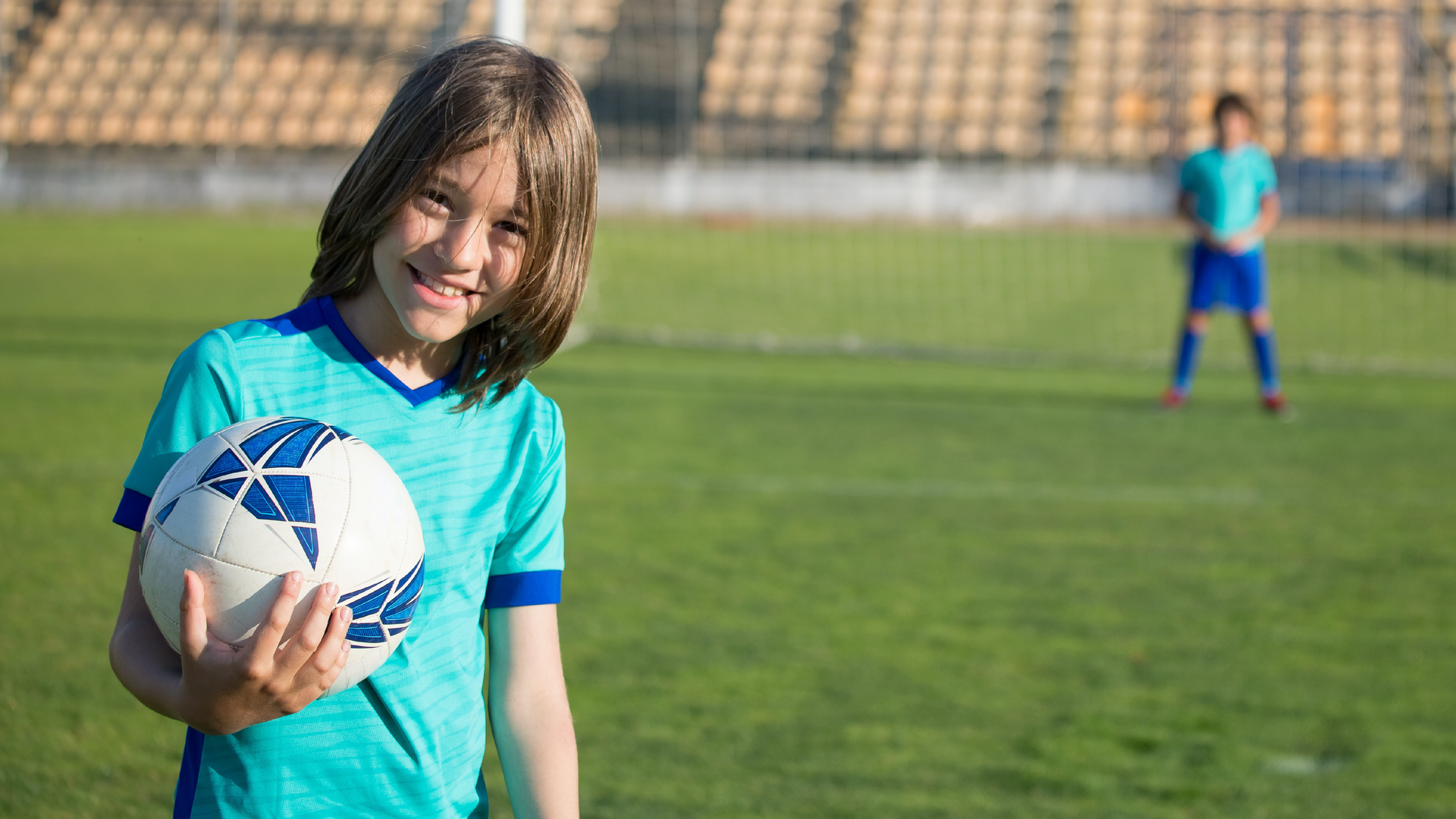 Zdjęcie przedstawia uśmiechniętego chłopca z długimi włosami. Chłopiec stoi na murawie piłkarskiej i trzyma w ręku piłkę. Jest ubrany w strój sportowy. W tle zdjęcie jest rozmyte i widać innego chłopca w stroju piłkarskim oraz trybuny.