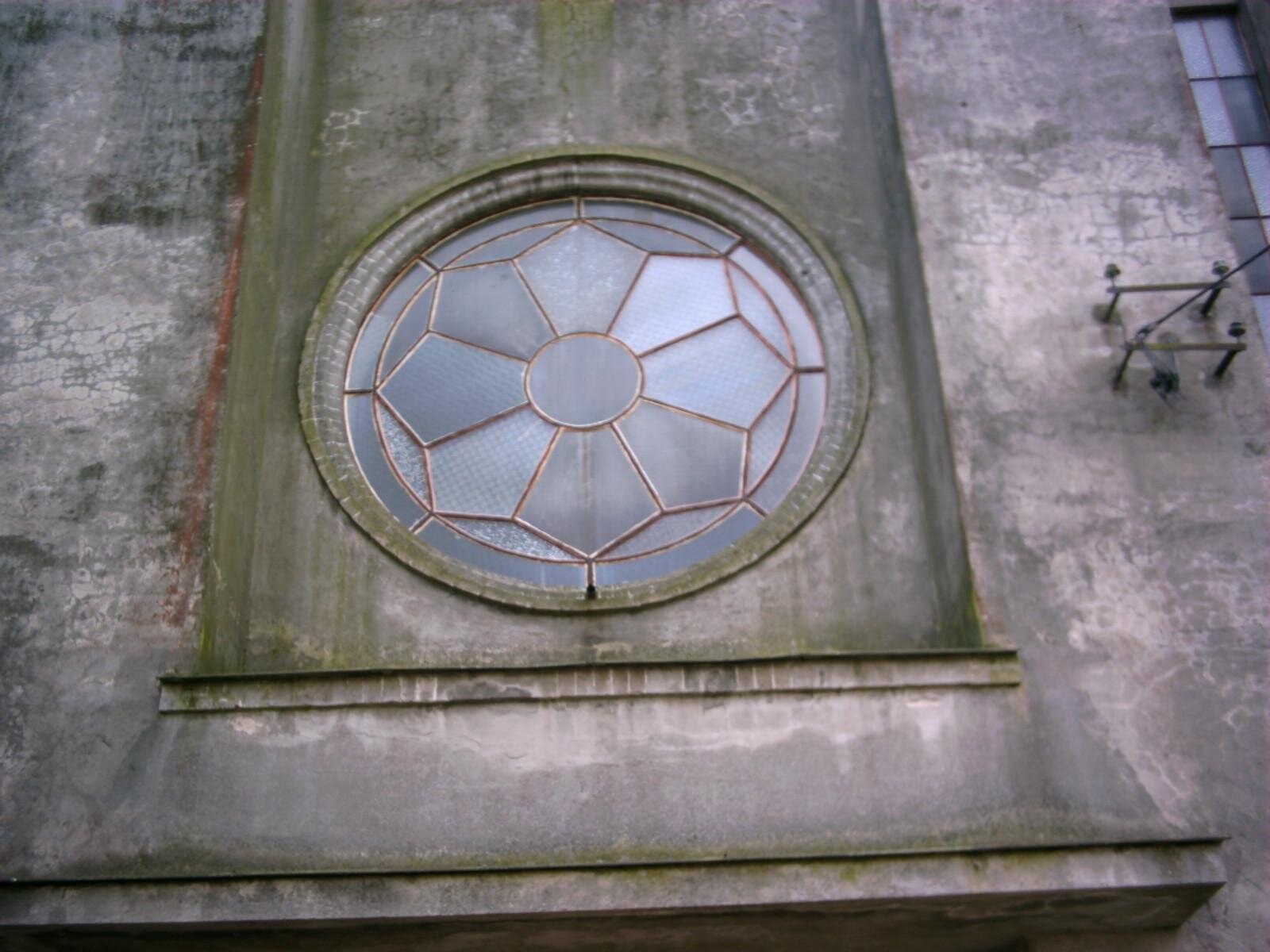 Zdjęcie przedstawia fragment szarej ściany, na której znajduje się okrągłe okno. Szyba okna jest podzielona pomarańczową farbą na części, które tworzą ośmiopłatkowego kwiata z okrągłym środkiem. Zdjęcie zostało wykonane w świetle dziennym.