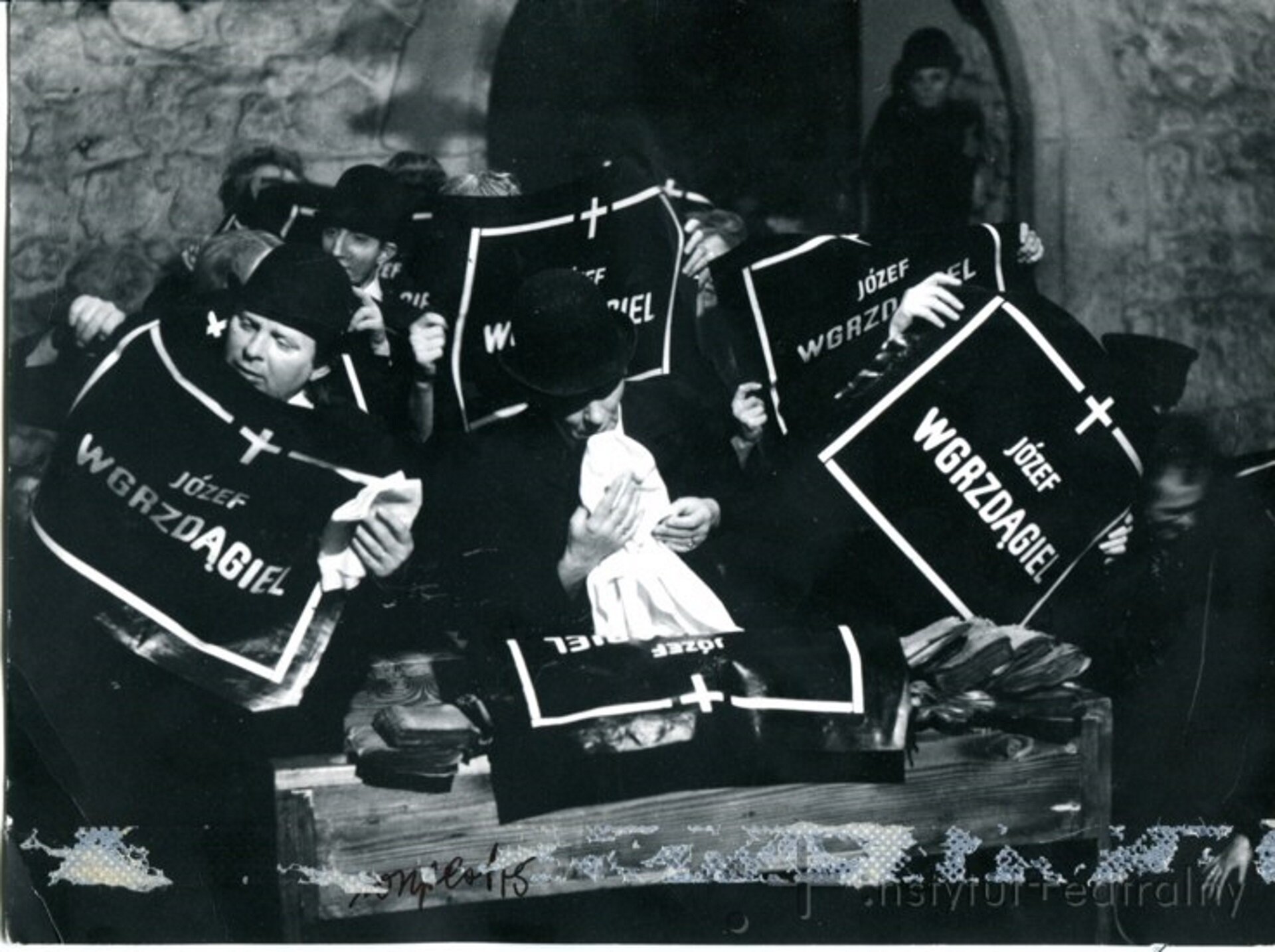 Ilustracja przedstawia czarno-białą fotografię, która prezentuje aktorów na scenie, ubrani są wszyscy na czarno, mają smutne miny. Trzymają w ręce nekrolog na czarnym papierze białą czcionką, tekst: Józef Wgrzdągiel. Aktorzy odgrywali wolę w spektaklu „Umarła klasa”.