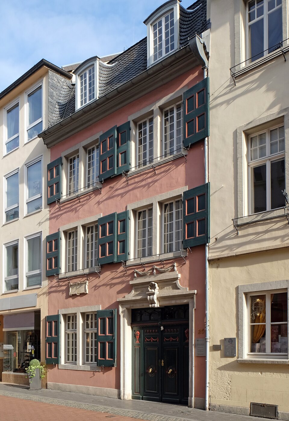 Ilustracja przedstawia dom, w którym urodził się Ludwig van Beethoven. Na fotografii widzimy fragment bliźniaczych kamienic. Dom ma elewację w odcieniu różu.