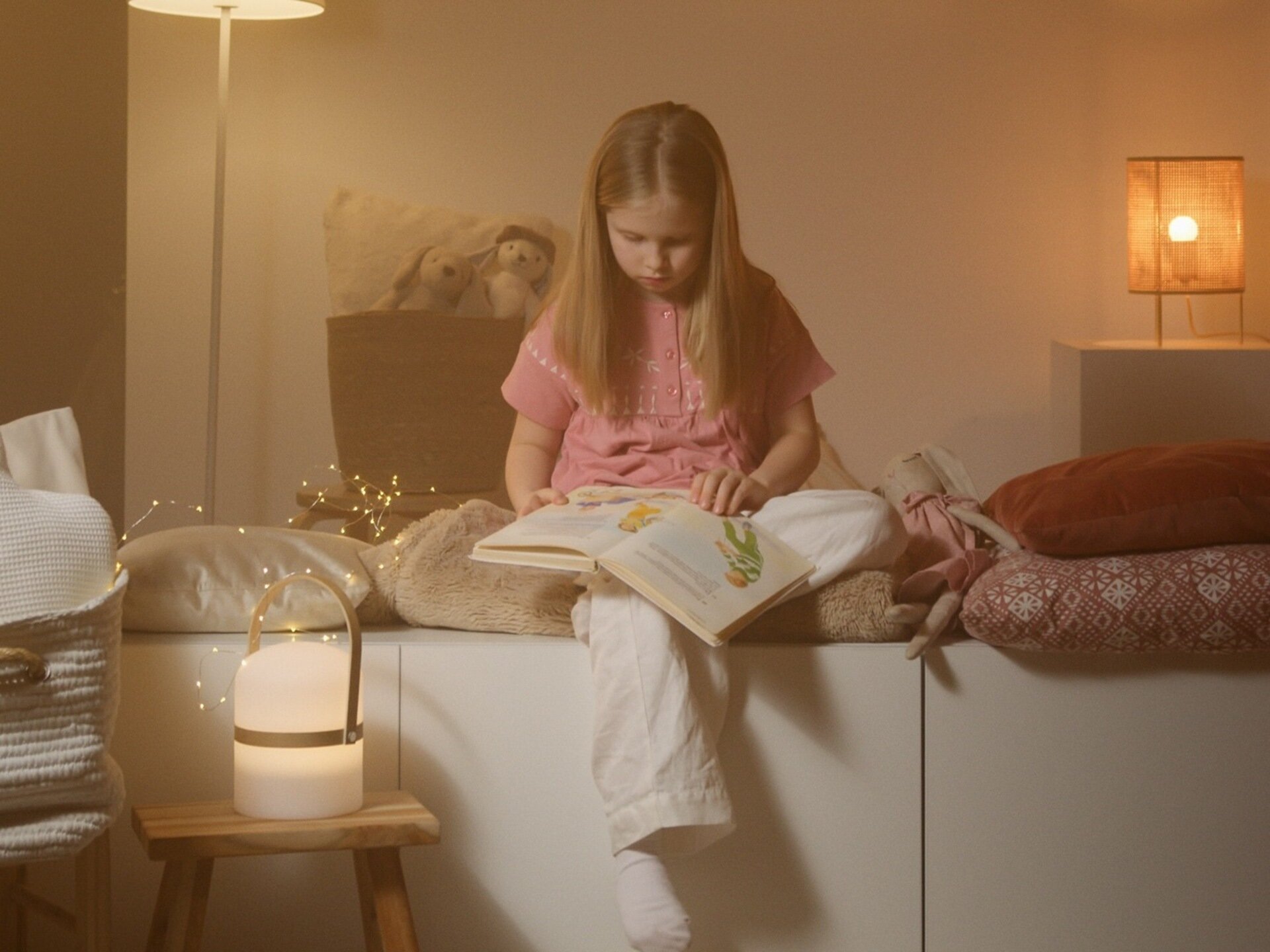Zdjęcie przedstawia dziewczynkę w wieku szkolnym siedzącą na oparciu sofy. Dziewczynka trzyma w dłoniach książkę i czyta ją.