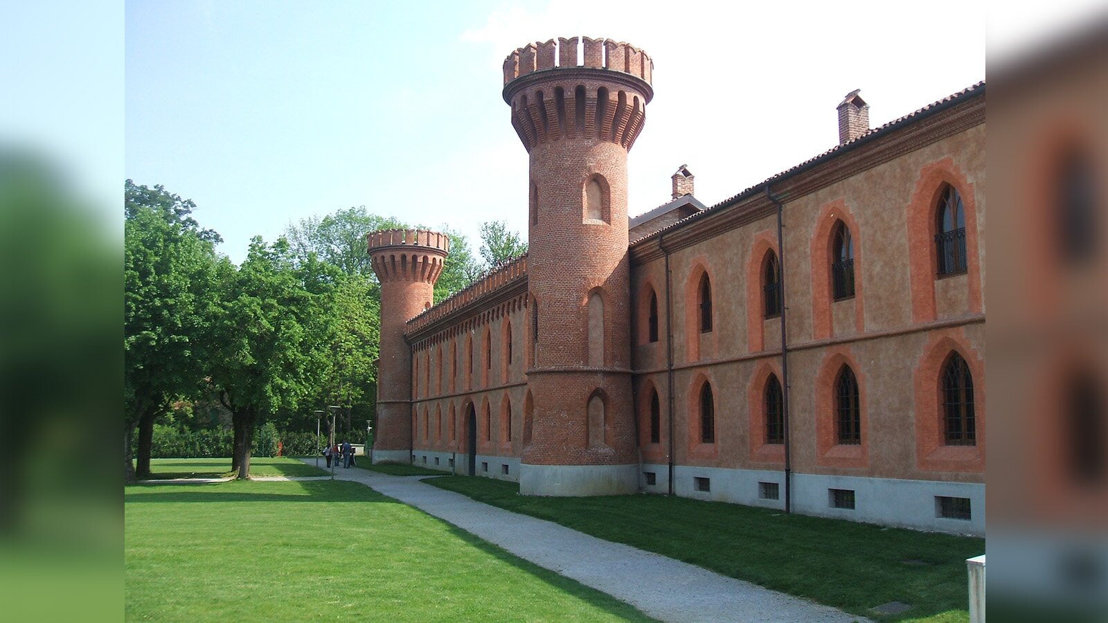 Zdjęcie przedstawia monumentalny, zabytkowy budynek z czerwonej cegły oraz przylegający do niego park. Budynek widoczny jest po prawej stronie. Zbudowany jest z dwóch wysokich pięter, z oknami zakończonymi u góry ostrymi łukami, oraz dwóch wysokich wież. Wzdłuż budynku biegnie pas zielonej trawy, szary chodnik, a następnie zielony teren z wysokimi drzewami. W głębi widoczne są postaci idące chodnikiem.
